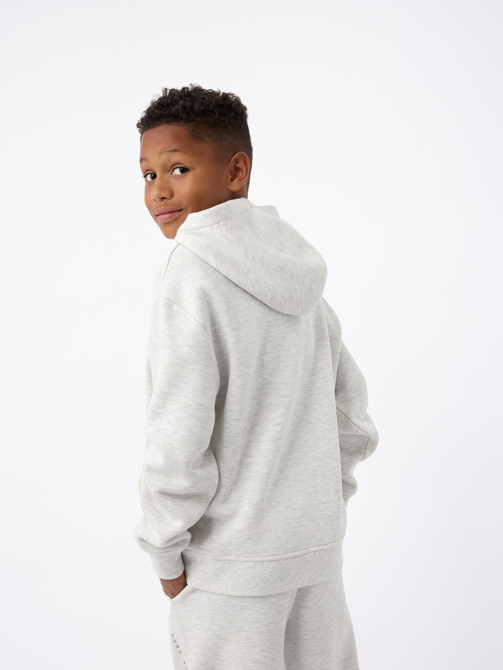 A young boy with short curly hair is wearing the Kid's CityScape Hoodie from Cozy Earth paired with matching sweatpants. He is looking over his shoulder with a slight smile against a plain white background. 