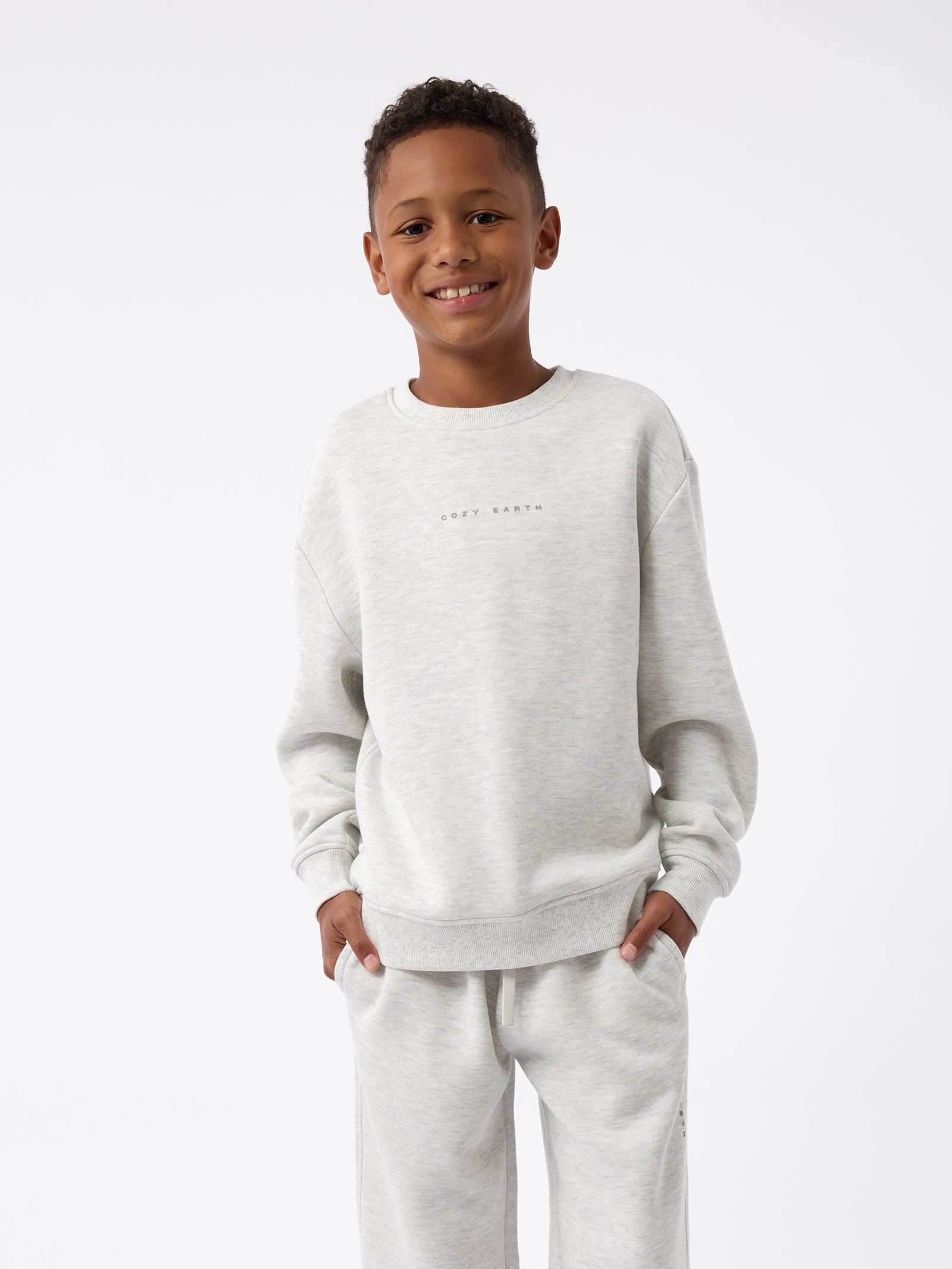 A smiling child stands wearing a light gray Kid's CityScape Sweatpant from Cozy Earth, with their hands in the pockets of their matching sweatshirt, against a plain white background. 