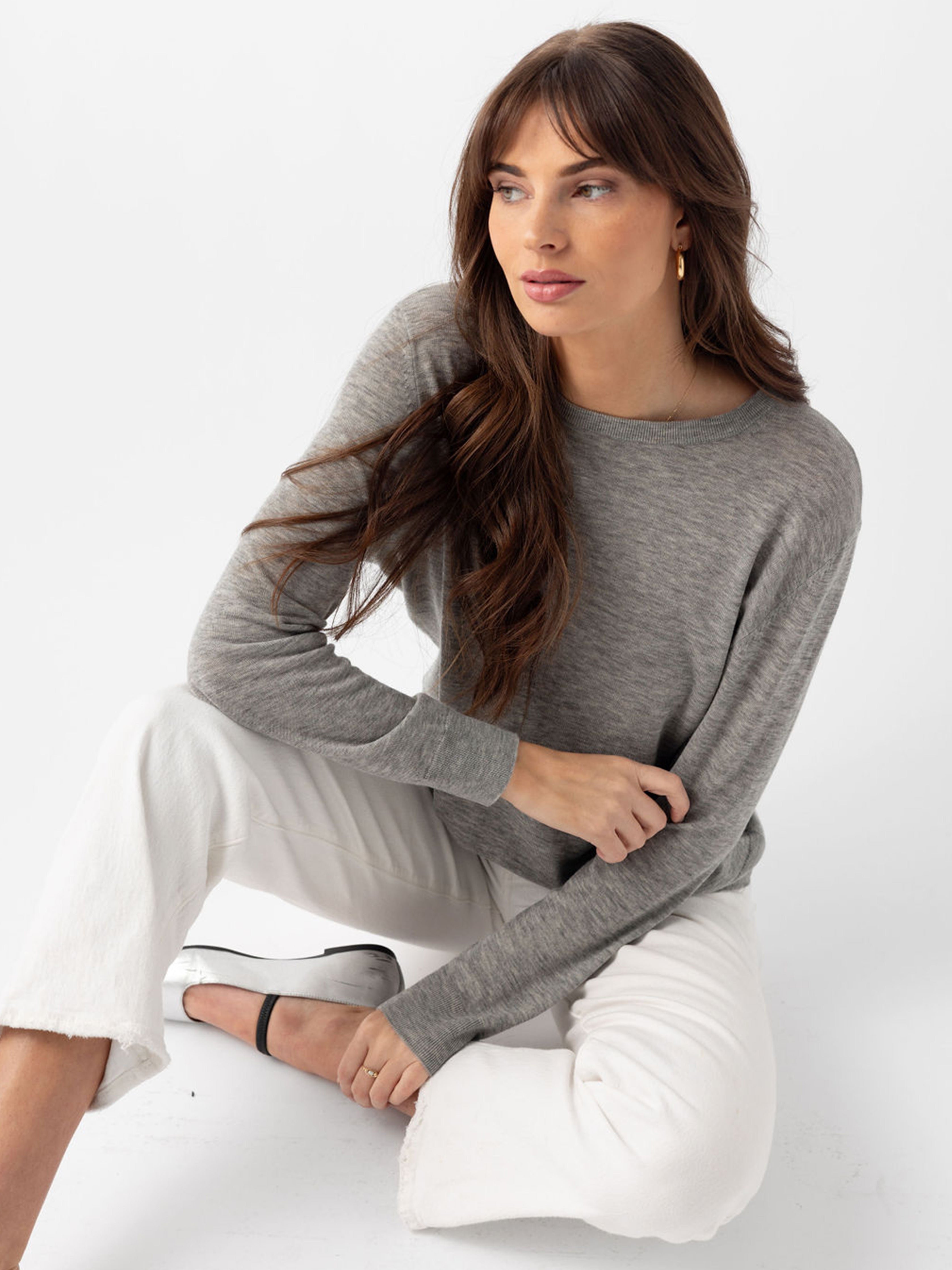 Woman sitting on the ground in heather grey airknit sweater and white jeans with white background 