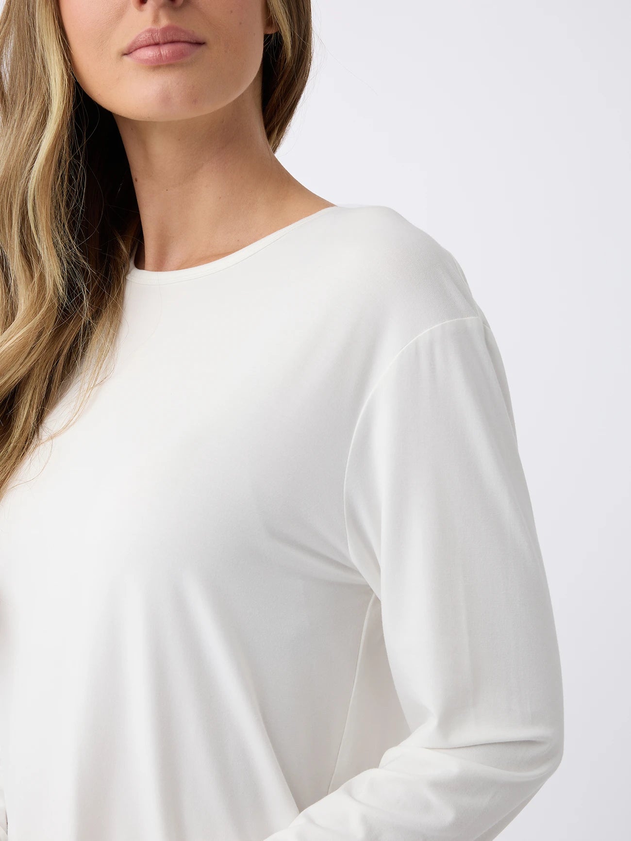 A woman with long, wavy hair is wearing a Women's Stretch Knit Bamboo Long Sleeve Lounge Tee by Cozy Earth. The photo captures the upper body from the side, showcasing the shirt's fabric and fit against a plain background. 