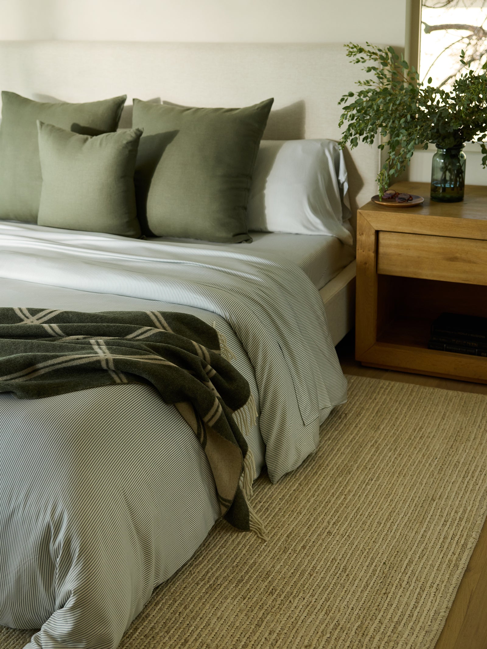 A cozy bedroom showcases a neatly made bed adorned with white and green Cozy Earth Bamboo Pillowcases. A green plaid blanket is draped over the edge, complementing the setup. Beside the bed sits a wooden nightstand holding a vase with a plant, while natural light filters through a nearby window. 