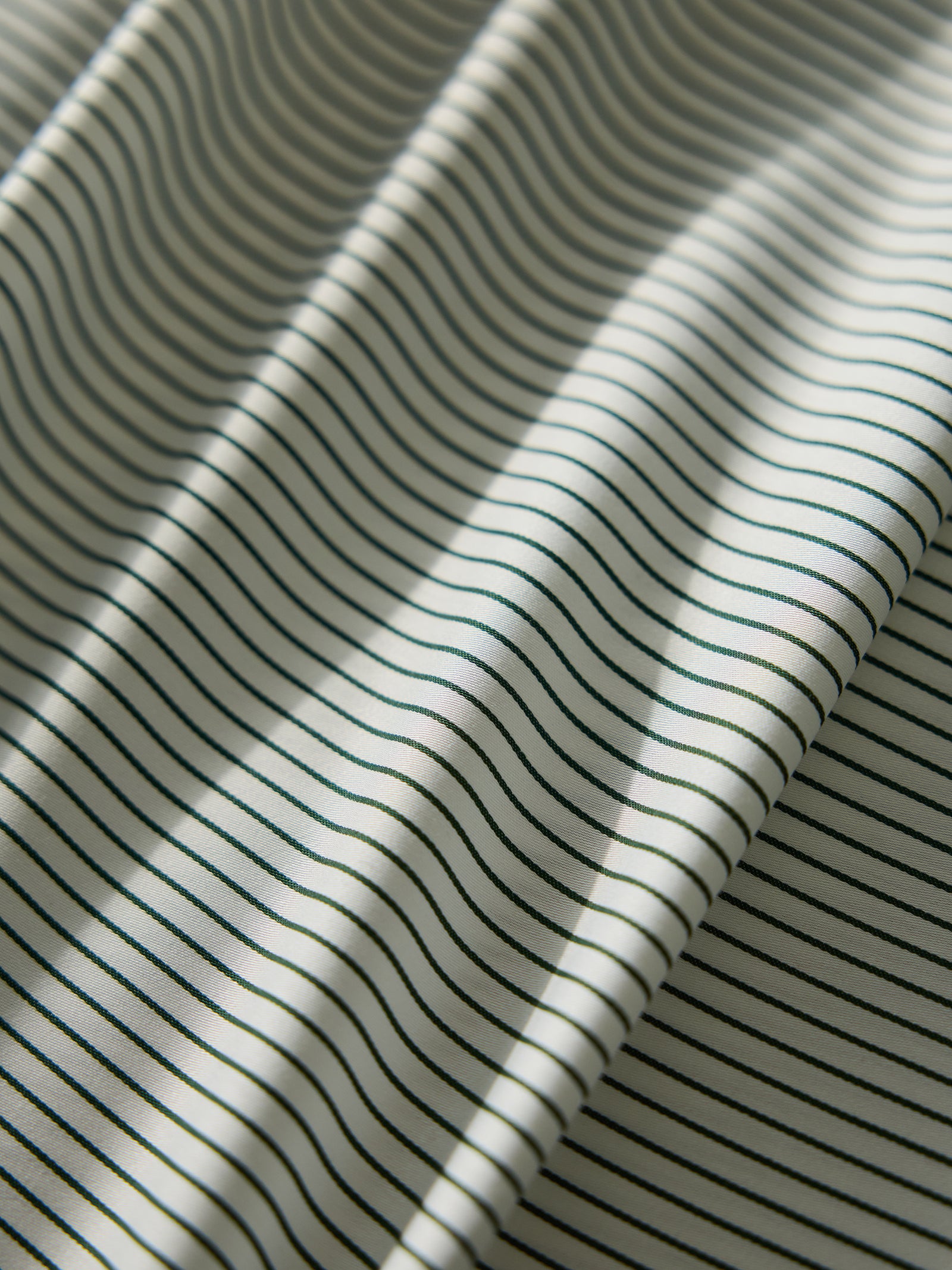 Close-up of the Bamboo Fitted Sheet by Cozy Earth, showcasing a pattern of thin, dark horizontal stripes on a light background. The sheet is softly folded, creating gentle waves that cast subtle shadows. 