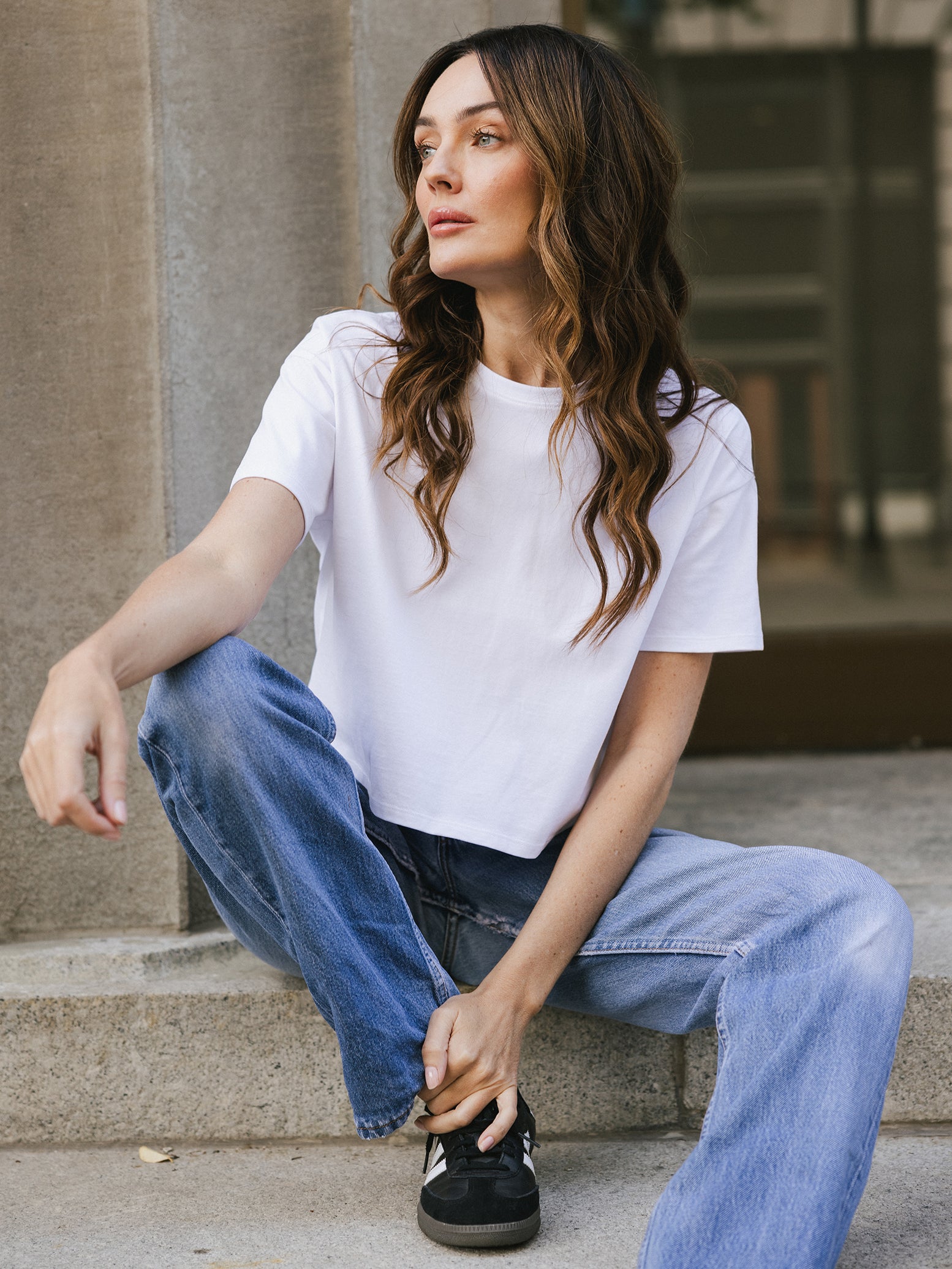 White All Day Cropped Tee. The photo of the All Day Cropped Tee is taken with a with a city style background and is worn by a woman. 