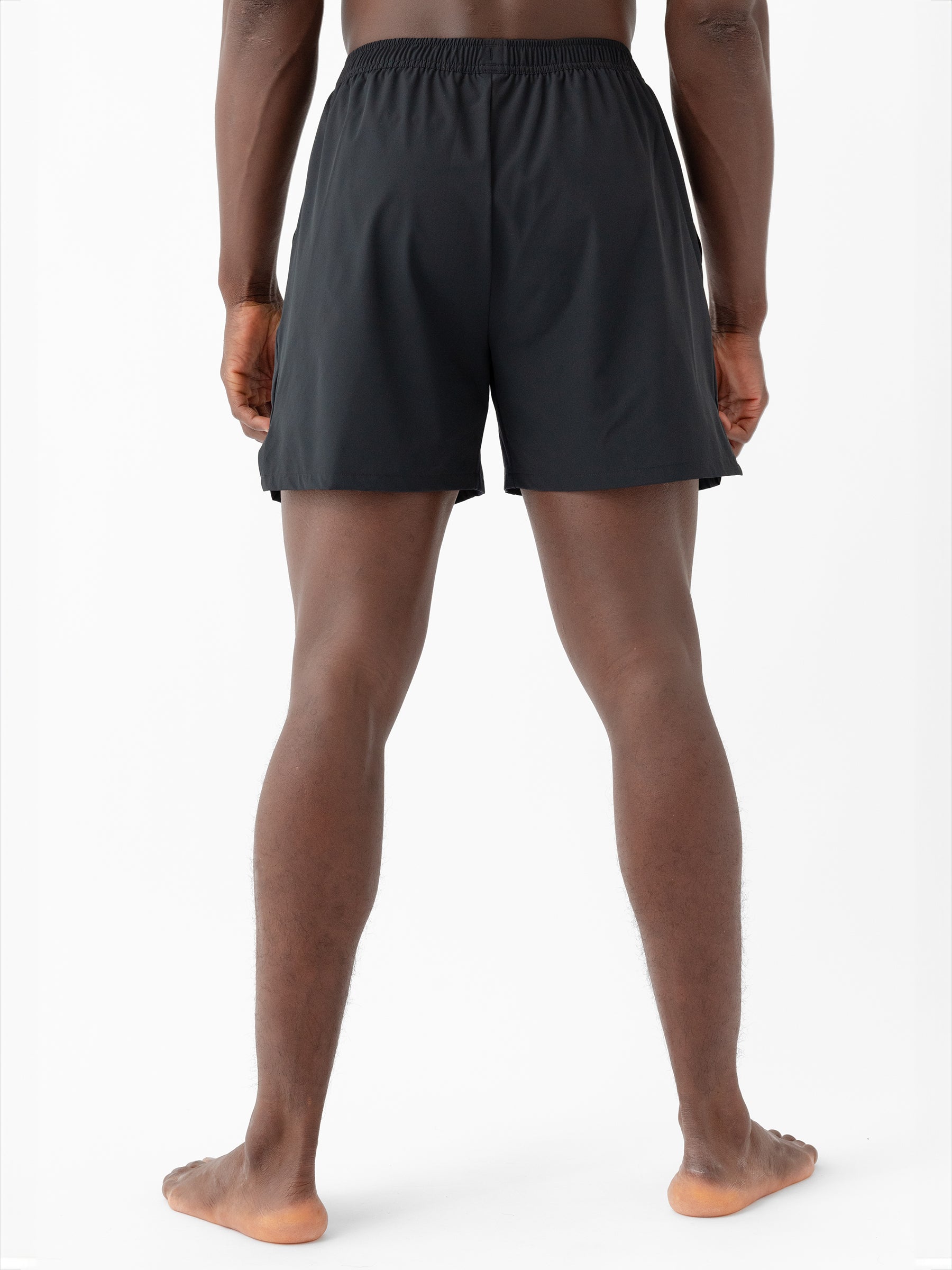 A man stands facing away from the camera, showcasing Cozy Earth's Men's Performance Sleep Short. He is barefoot, and the photo is taken from the back, highlighting the shorts' length and fit against a plain white background. |Color:Jet Black