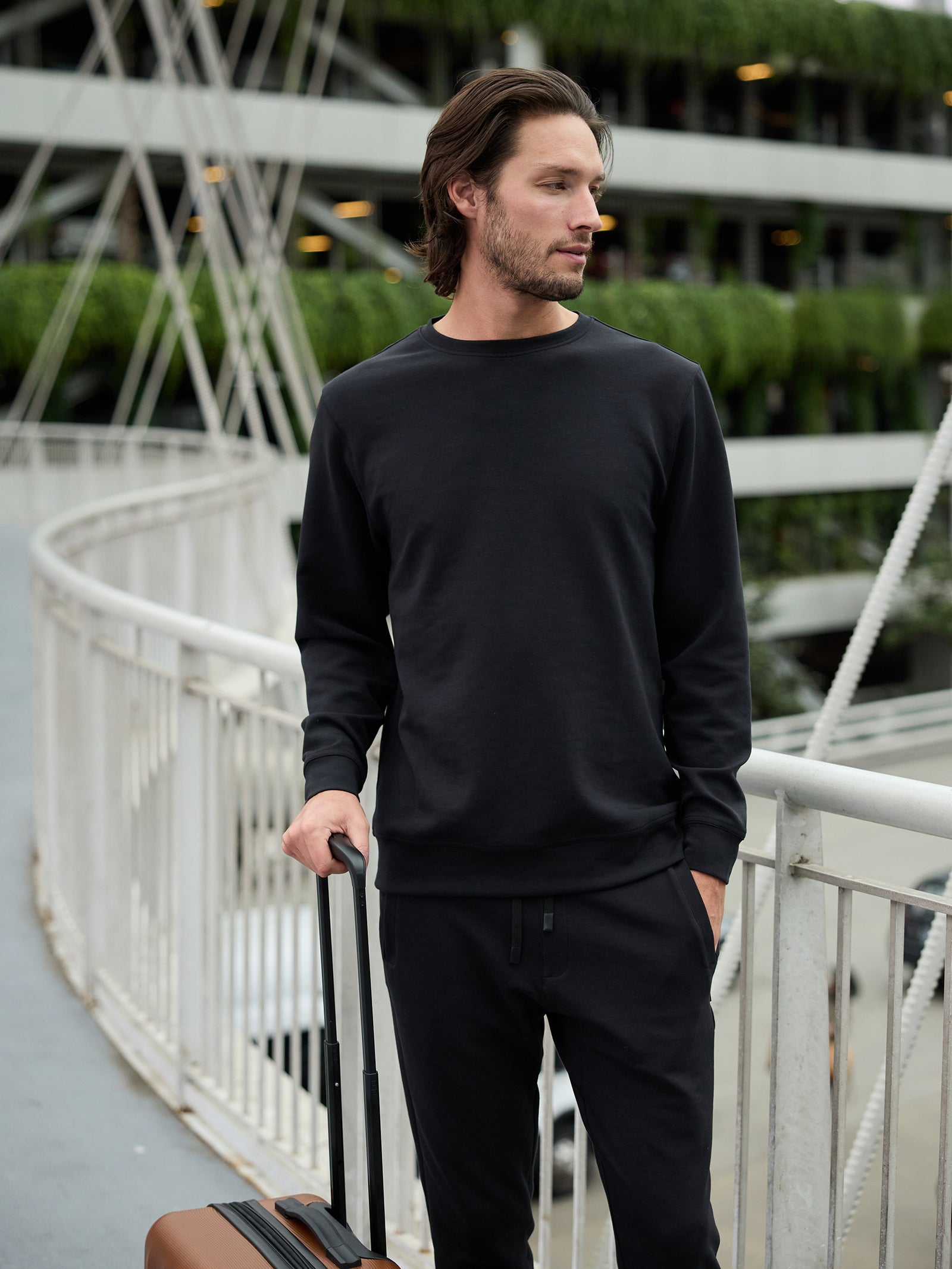 A man with shoulder-length hair and facial hair stands on a curved pedestrian bridge. He is wearing a Cozy Earth Men's StretchTech Crewneck sweater and black pants, holding a suitcase with his right hand. In the background, there is a multi-story building adorned with green plants on its balconies. 