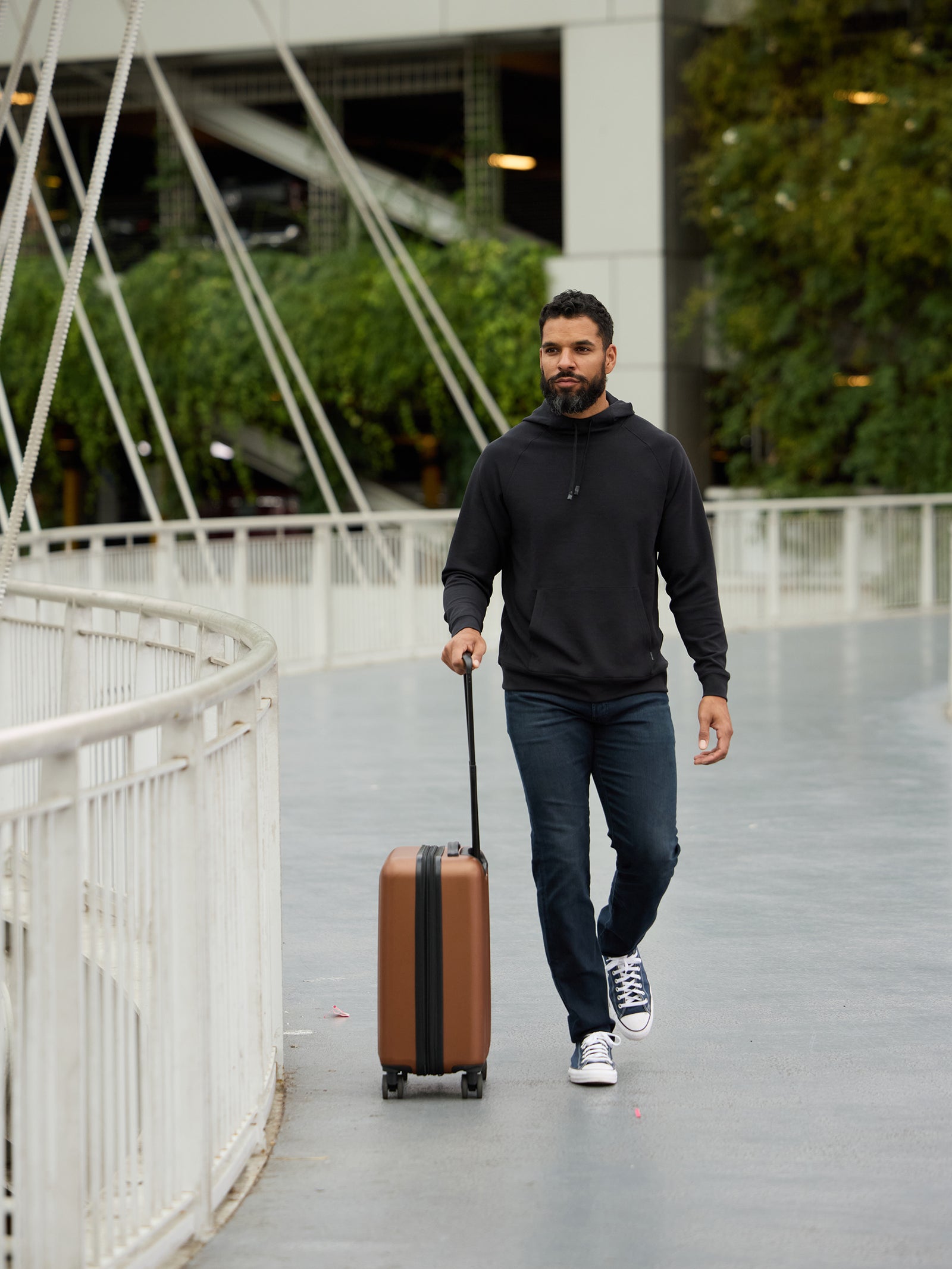 A bearded man in a Cozy Earth Men's StretchTech Hoodie and blue jeans walks along an outdoor walkway, pulling a brown wheeled suitcase behind him. He is on a modern bridge with white railings, and green trees with a building are visible in the background. 