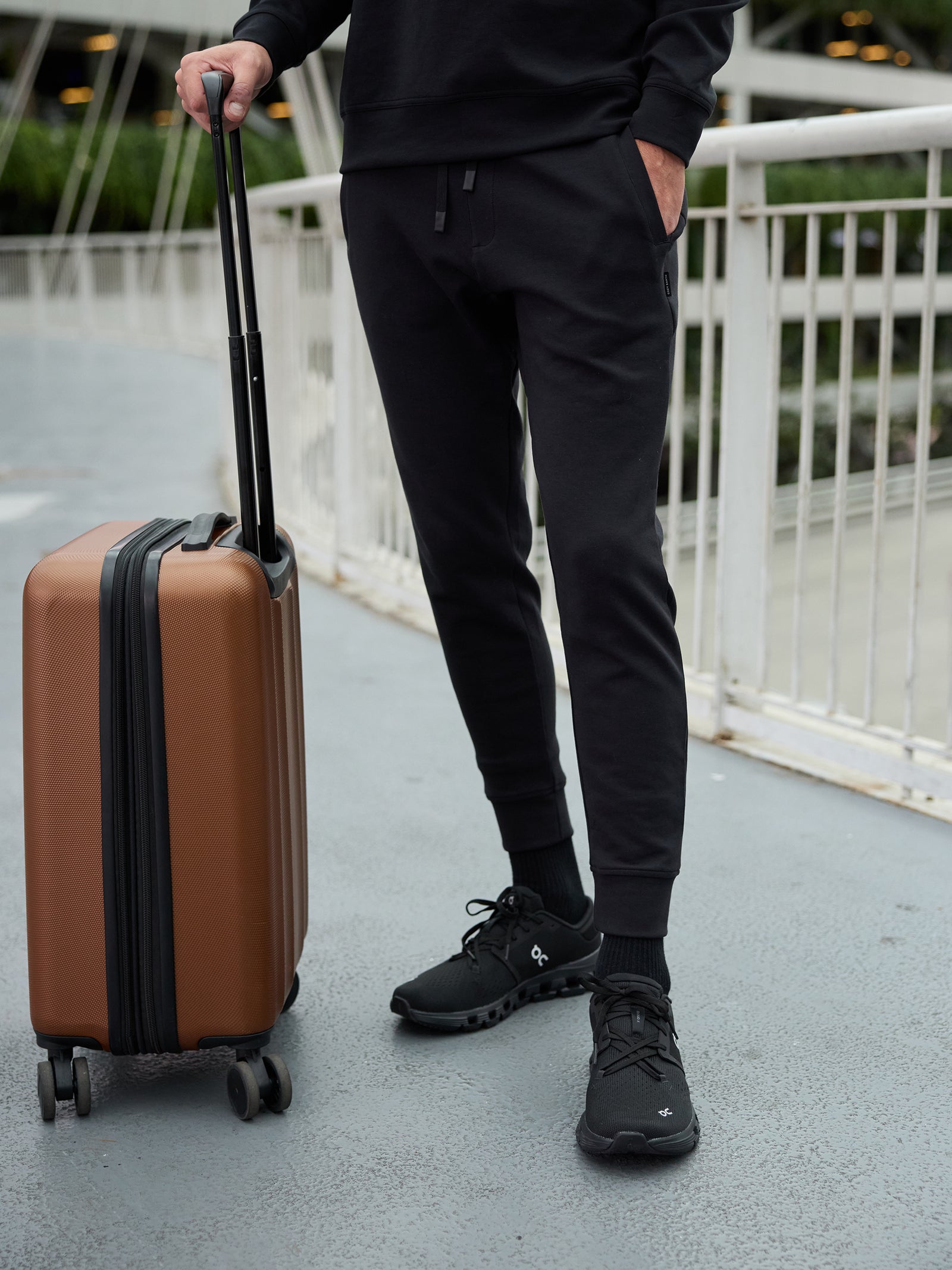 A person dressed in black casual attire, including a long-sleeve top, Men's StretchTech Jogger pants from Cozy Earth, and sneakers, stands on a bridge. They are holding the handle of a brown wheeled suitcase. The background features a white railing and green trees. 