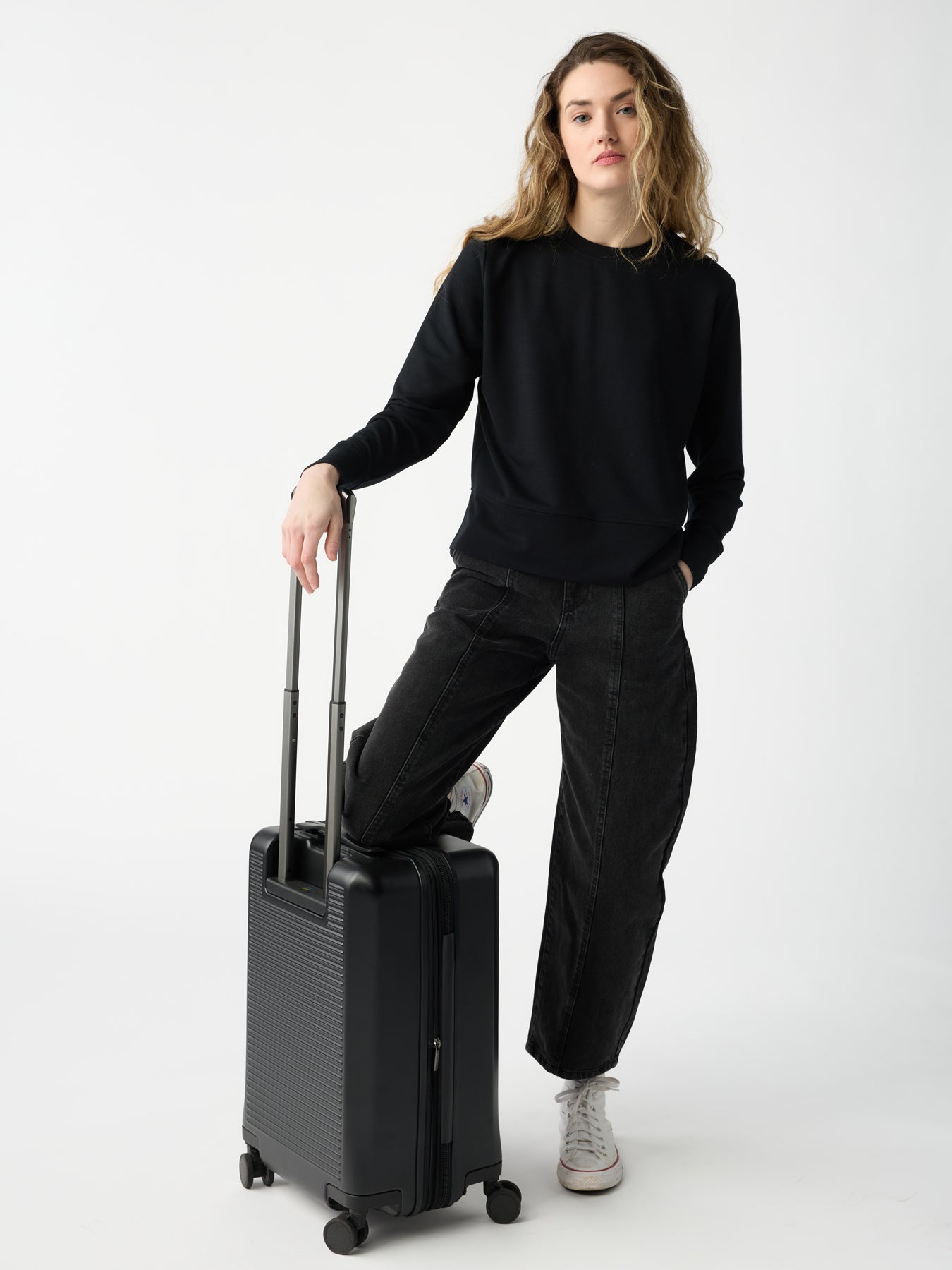 A person with long hair wearing a Cozy Earth Women's StudioLite Crewneck and dark jeans stands on a white background, resting one hand on the handle of a black rolling suitcase. 