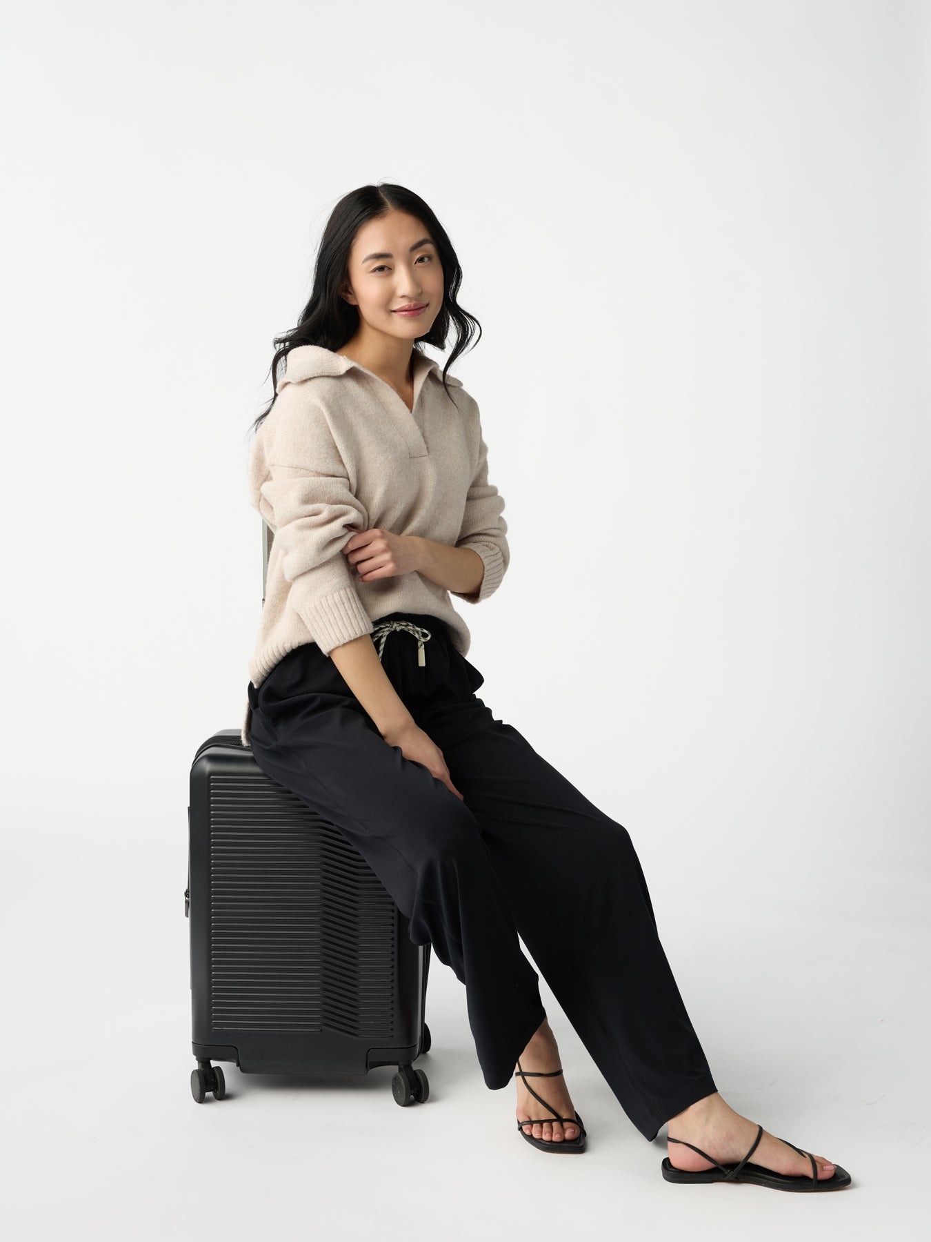 A woman with long black hair sits on a black suitcase against a white background, wearing a beige sweater, Cozy Earth's Women's Studio Wide Leg Pant in black, and black sandals. She appears relaxed and confident. 