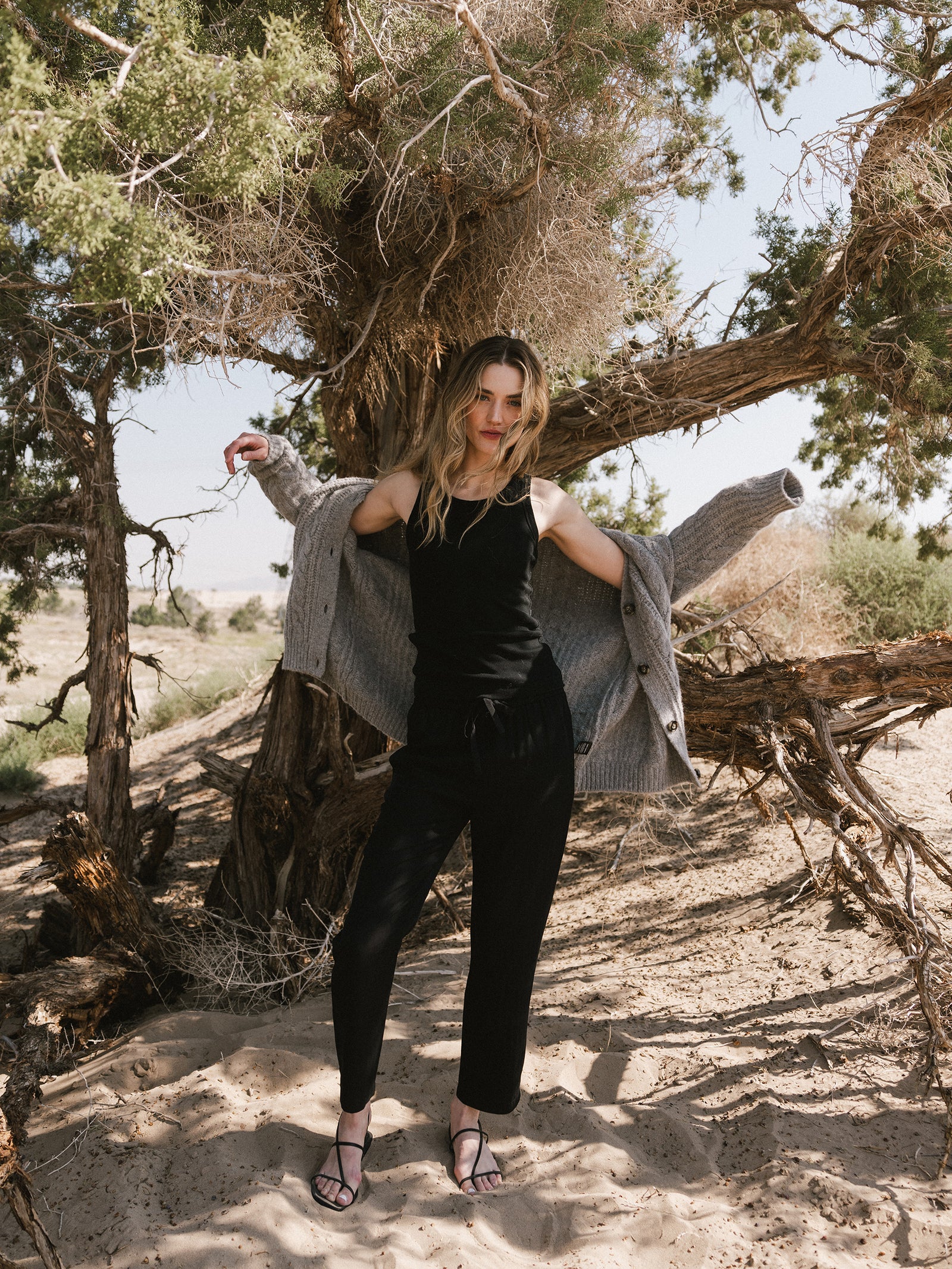 A person stands in a sandy, desert-like area with sparse, rugged trees. They wear a black tank top, Cozy Earth's Women's Sunset Cropped Pant, and sandals, with a gray cardigan draped over their shoulders. Their long hair is loose, and they display a relaxed yet thoughtful expression. 