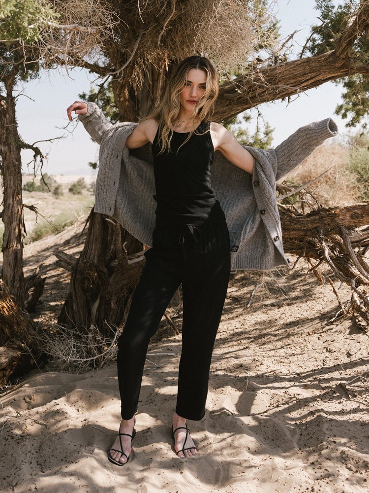 A woman stands on sandy ground under a large tree, wearing Cozy Earth’s Women’s Sunset Cropped Pant, a black tank top, sandals, and a gray cardigan draped over her shoulders as sunlight filters through the branches. 