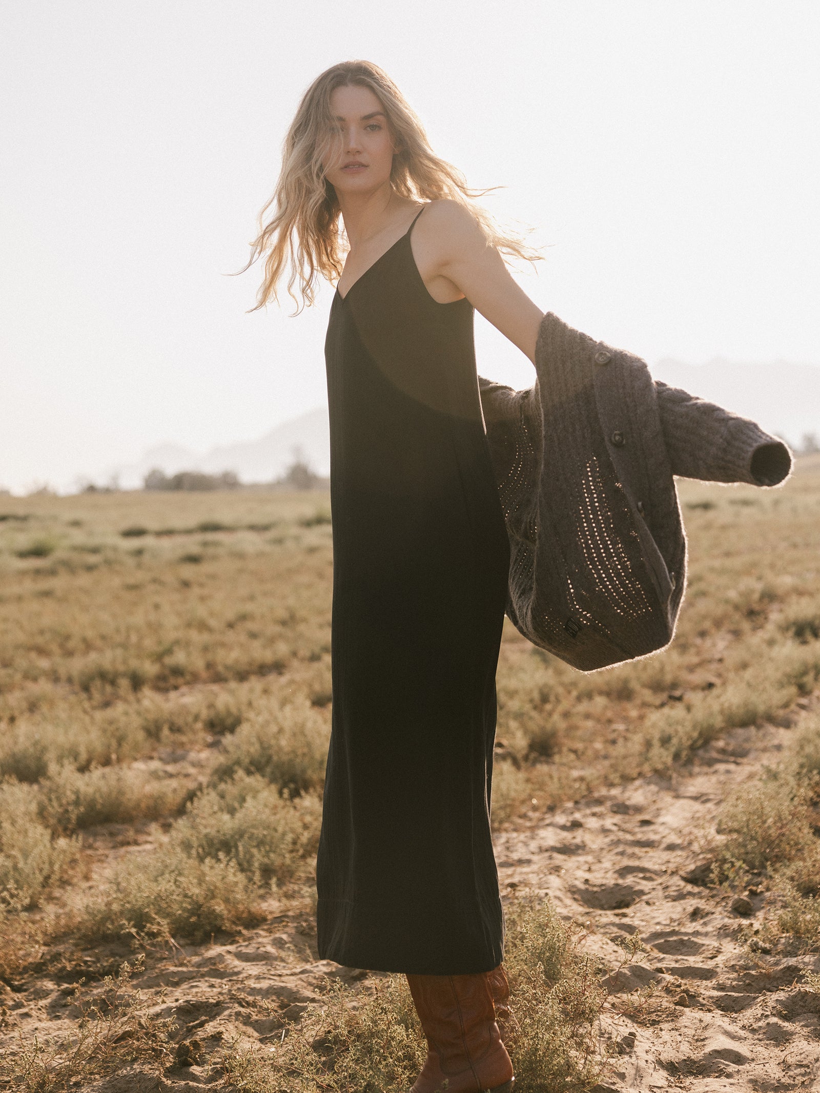 A woman with long wavy hair stands in a field, wearing the Women's Sunset Slip Dress by Cozy Earth and holding a gray knit cardigan. She gazes into the distance with a serene expression as the sun backlights her, casting a warm glow over the scene. 