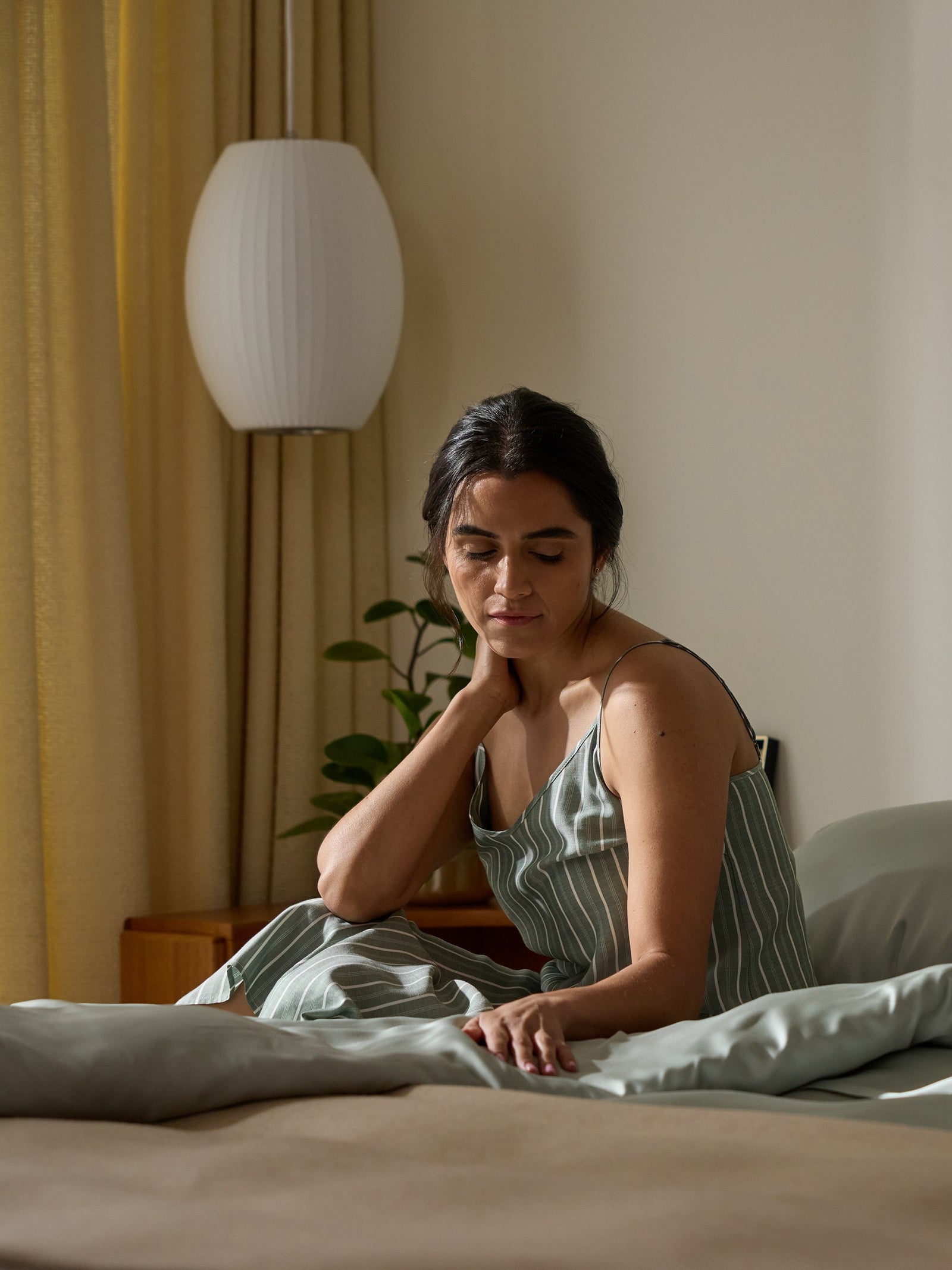 A woman sits thoughtfully on a bed adorned with a Cozy Earth Bamboo Duvet Cover in a softly lit room. Wearing a striped dress, she touches her neck gently. A lamp and small plant sit nearby against drawn curtains. 