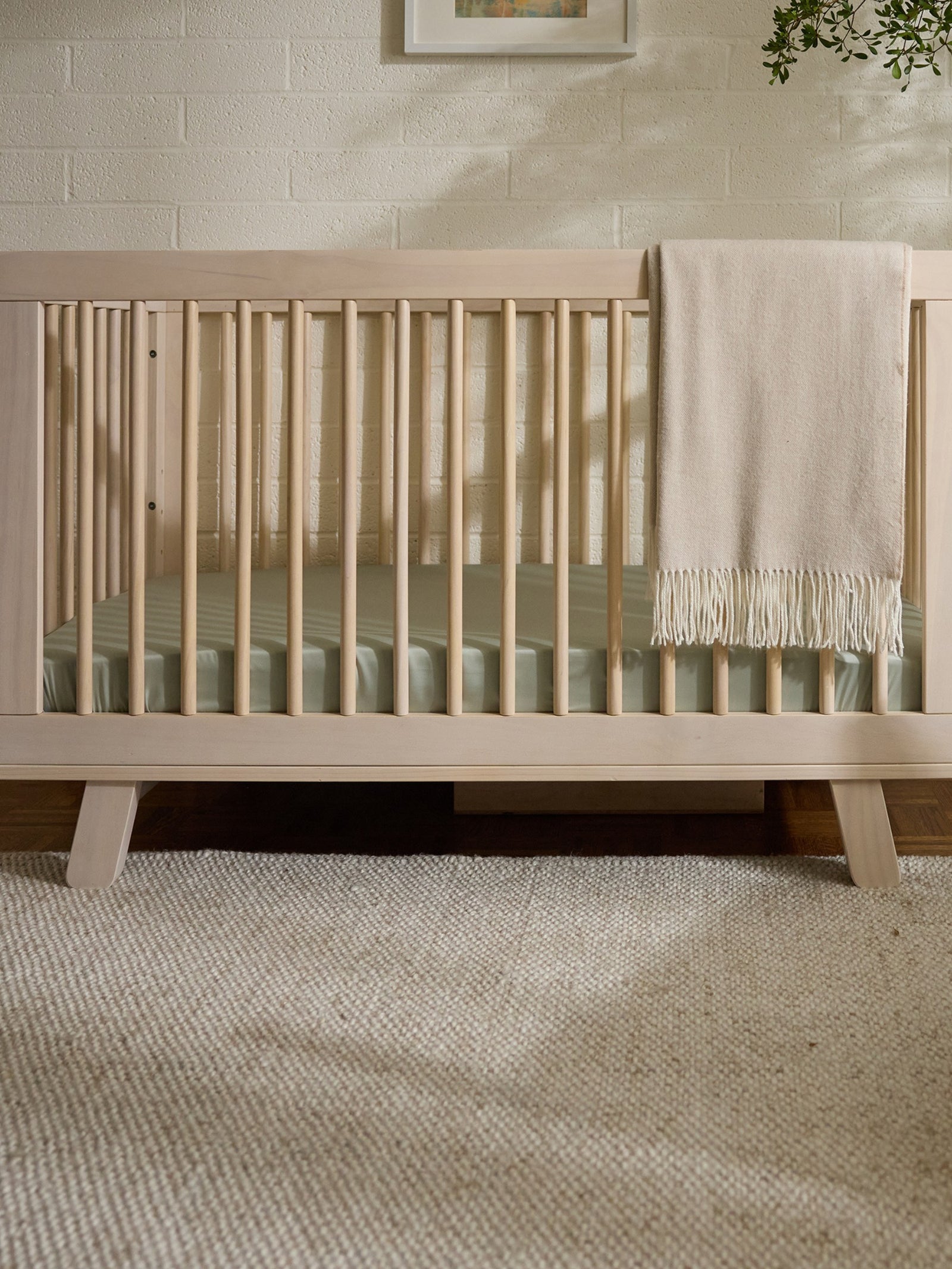 A wooden baby crib with vertical bars, featuring a Cozy Earth Crib Sheet by Cozy Earth, is draped with a beige blanket and set on a textured rug. The light-colored wall behind displays part of a framed picture, while a plant partially hangs into view. 