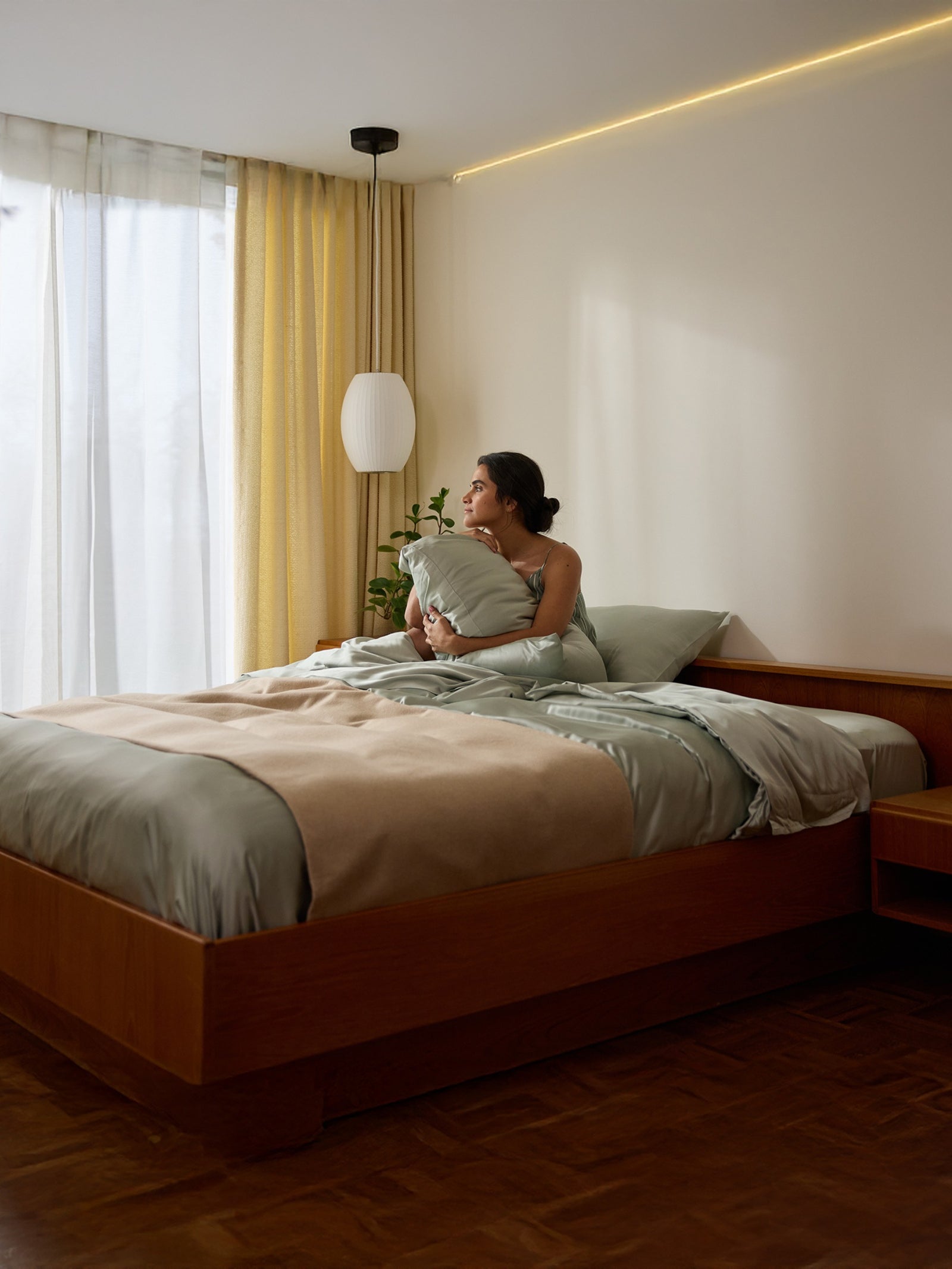 A person sits on a bed with Cozy Earth's Bamboo Pillowcases, smiling while gazing out the window. The room boasts light curtains, a potted plant, and soft ceiling lighting. The bed is adorned with light bedding. 