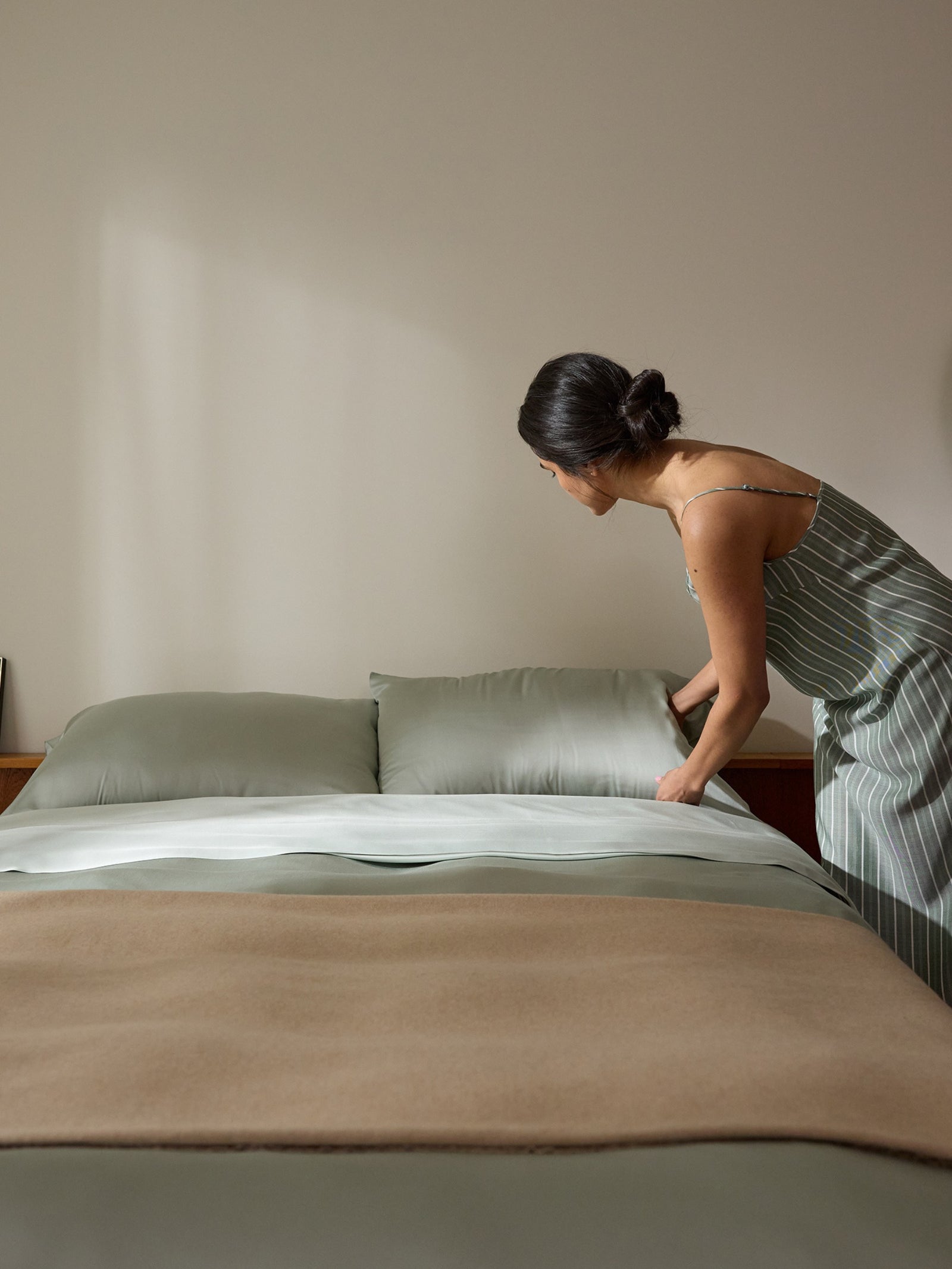 A person in a striped dress makes a bed with Cozy Earth's Bamboo Sheet Set, featuring green sheets and pillows. The room's decor is neutral and minimalist, highlighted by a sunlit wall in the background. 