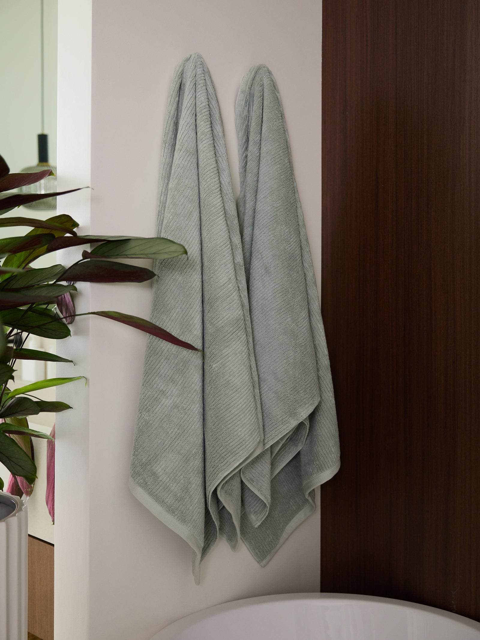 Two Cozy Earth Ribbed Terry Bath Towels in light gray hang side by side on a white wall in a bathroom. A dark wooden panel and a modern bathtub are partially visible nearby. A green leafy plant adds a touch of natural decor to the space. 