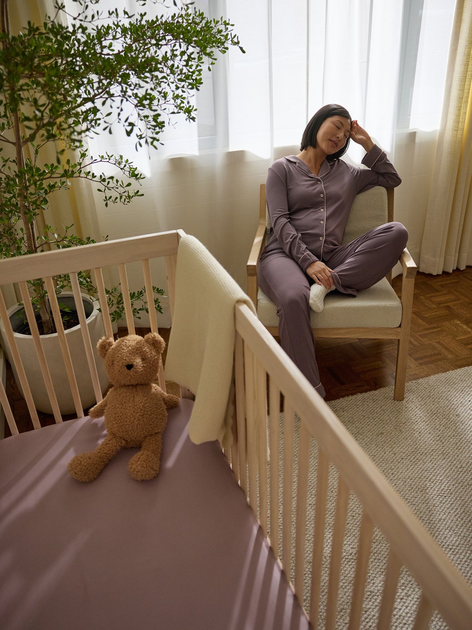 A woman in pajamas sits contentedly on a chair by a window in a nursery. A teddy bear rests on a Cozy Earth Crib Sheet from Cozy Earth in the nearby wooden crib. A potted plant adds greenery to the serene, well-lit room. 