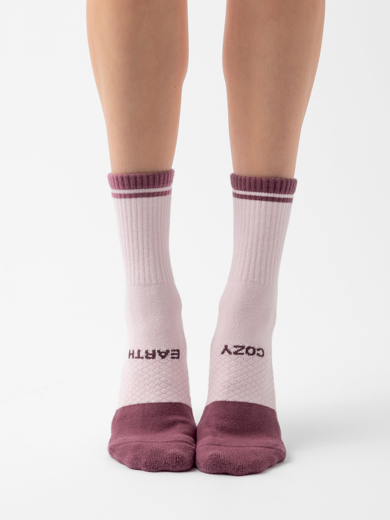 A pair of feet wearing Essential Calf Socks from Cozy Earth, featuring white socks with maroon toes, heels, and cuffs, standing on a white surface. The left sock displays the word "EARTH," while the right sock features the word "COZY. 