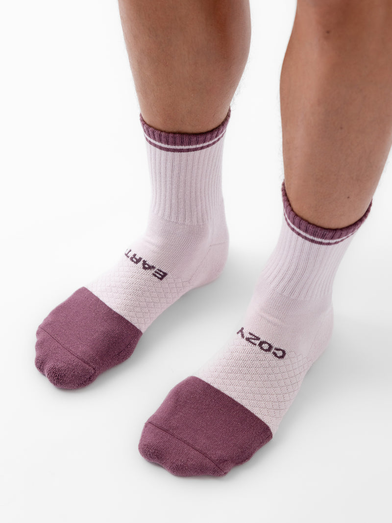 A close-up view of a person's lower legs and feet wearing a pair of Cozy Earth's Essential Quarter Socks from the 2-Pack collection. The white socks feature purple toes, heels, and stripes around the top, along with the words "COZY" and "EARTH" printed on the soles. The person stands on a white surface.