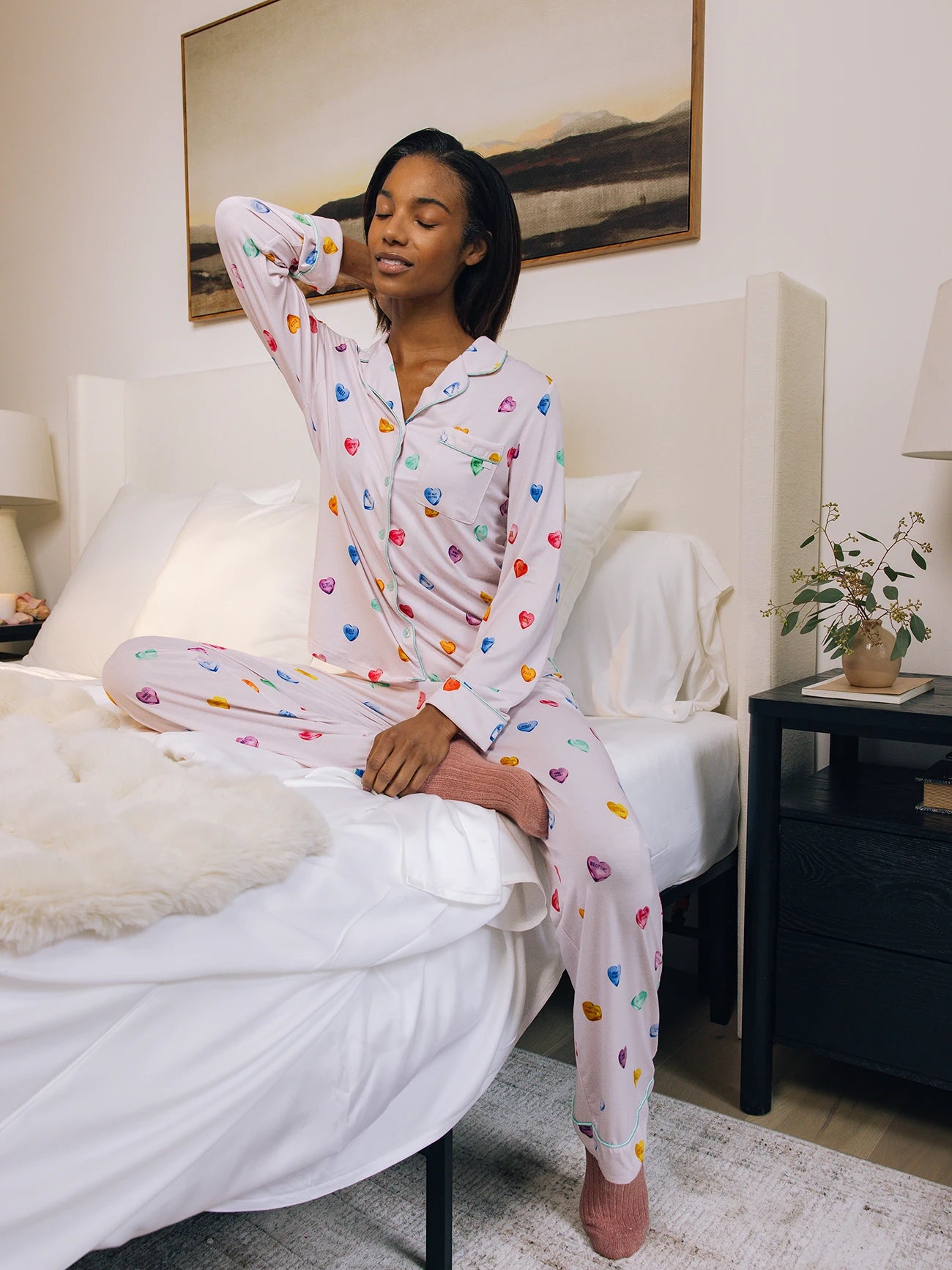 A woman in Cozy Earth's Women's Stretch-Knit Long Sleeve Bamboo Pajama Set stretches on a bed. The cozy room features a nightstand with plants, a lamp, and a wall painting. 