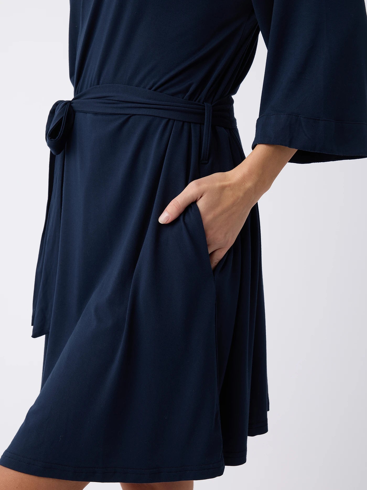 A person is wearing a Women's Stretch-Knit Bamboo Kimono Robe in dark blue from Cozy Earth. The knee-length robe features a waist tie and has the right hand tucked into its side pocket, all set against a plain white background. 