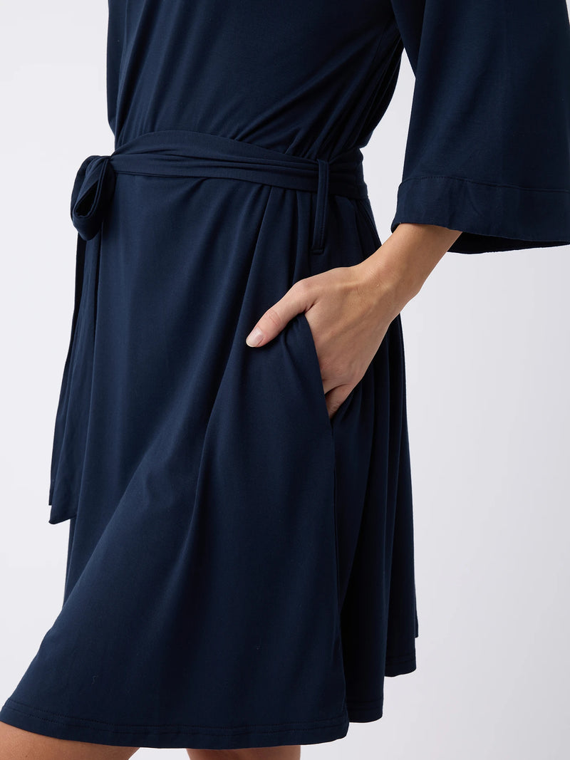 A person is wearing a Women's Stretch-Knit Bamboo Kimono Robe in dark blue from Cozy Earth. The knee-length robe features a waist tie and has the right hand tucked into its side pocket, all set against a plain white background.