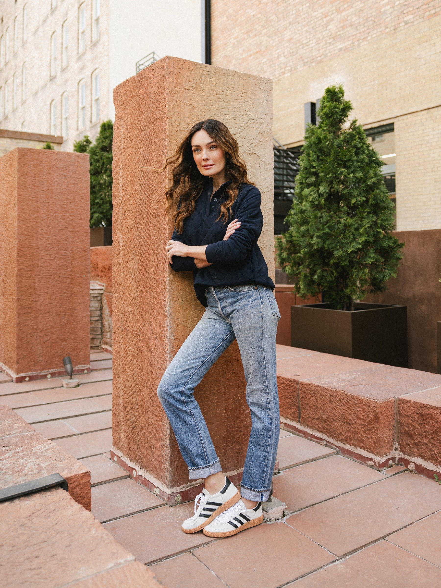 A woman with long, wavy hair stands leaning against a tall, textured column in an outdoor urban setting. She is wearing a navy Women's Ultra-Soft Bamboo Quilted Snap Pullover from Cozy Earth, light blue jeans, and white Adidas sneakers with black stripes. Green shrubs in planters decorate the background. 