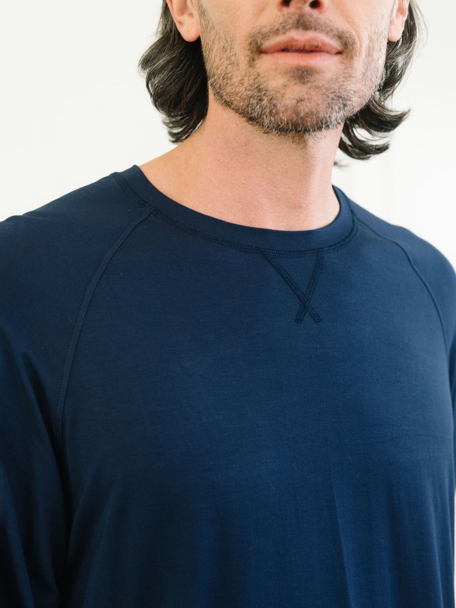 A man with shoulder-length hair and a beard is wearing a dark blue top from the Men's Stretch-Knit Long Sleeve Bamboo Pajama Set by Cozy Earth. The image is cropped to show only the lower half of his face and upper torso. 