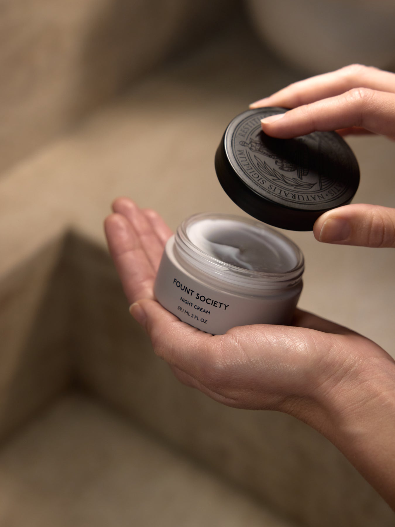 Two hands hold an open jar of Cozy Earth Night Cream against a neutral background. One hand removes the black lid while the other supports the jar, showing the cream inside ready for use. 