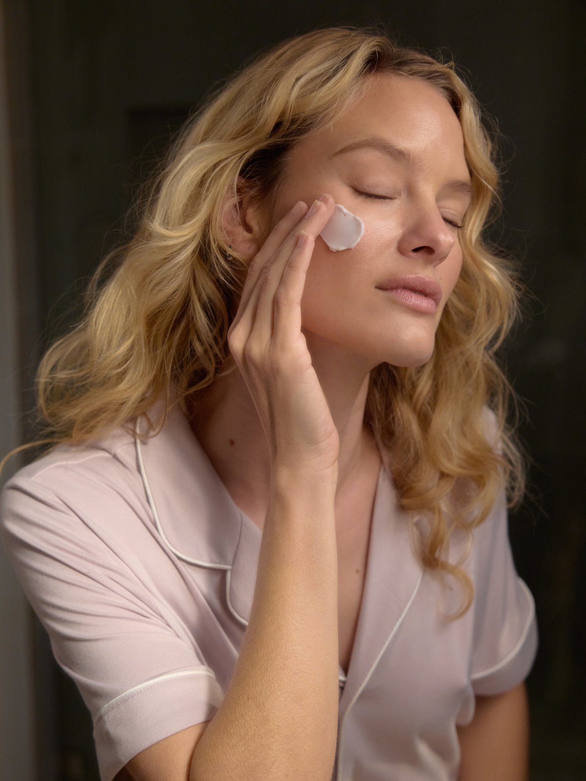 A person with long, wavy blonde hair applies a small amount of Cozy Earth's Day-to-Night Essentials facial cream to their cheek. Wearing a pale pink top with white piping, they have their eyes closed and appear relaxed against a softly blurred background.