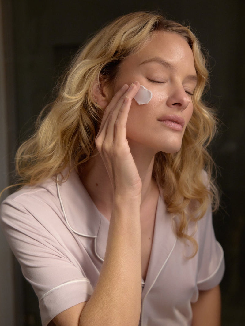 A woman with long blonde hair applies Cozy Earth's Night Cream to her cheek. She's wearing a light, short-sleeved collared top and appears relaxed with closed eyes in a softly lit room.