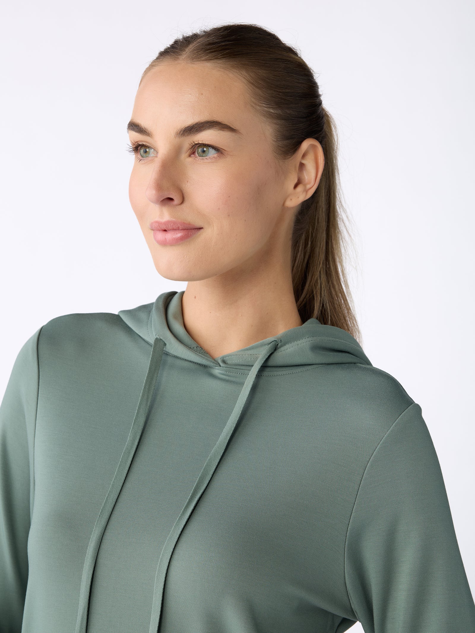 A person with long hair is looking to the side while wearing a Women's Ultra-Soft Bamboo Hoodie in sage green from Cozy Earth. The background is plain white. 