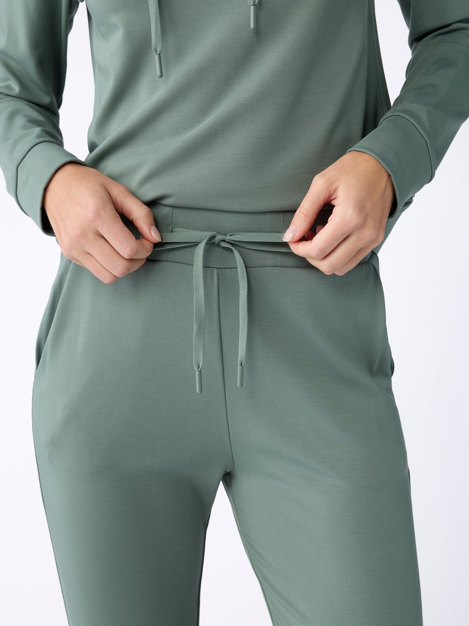 A person wearing the Women's Bamboo Jogger Pant TALL in green from Cozy Earth adjusts the drawstring on the pants. The image is focused on the torso and hands, highlighting the comfortable, casual outfit. 
