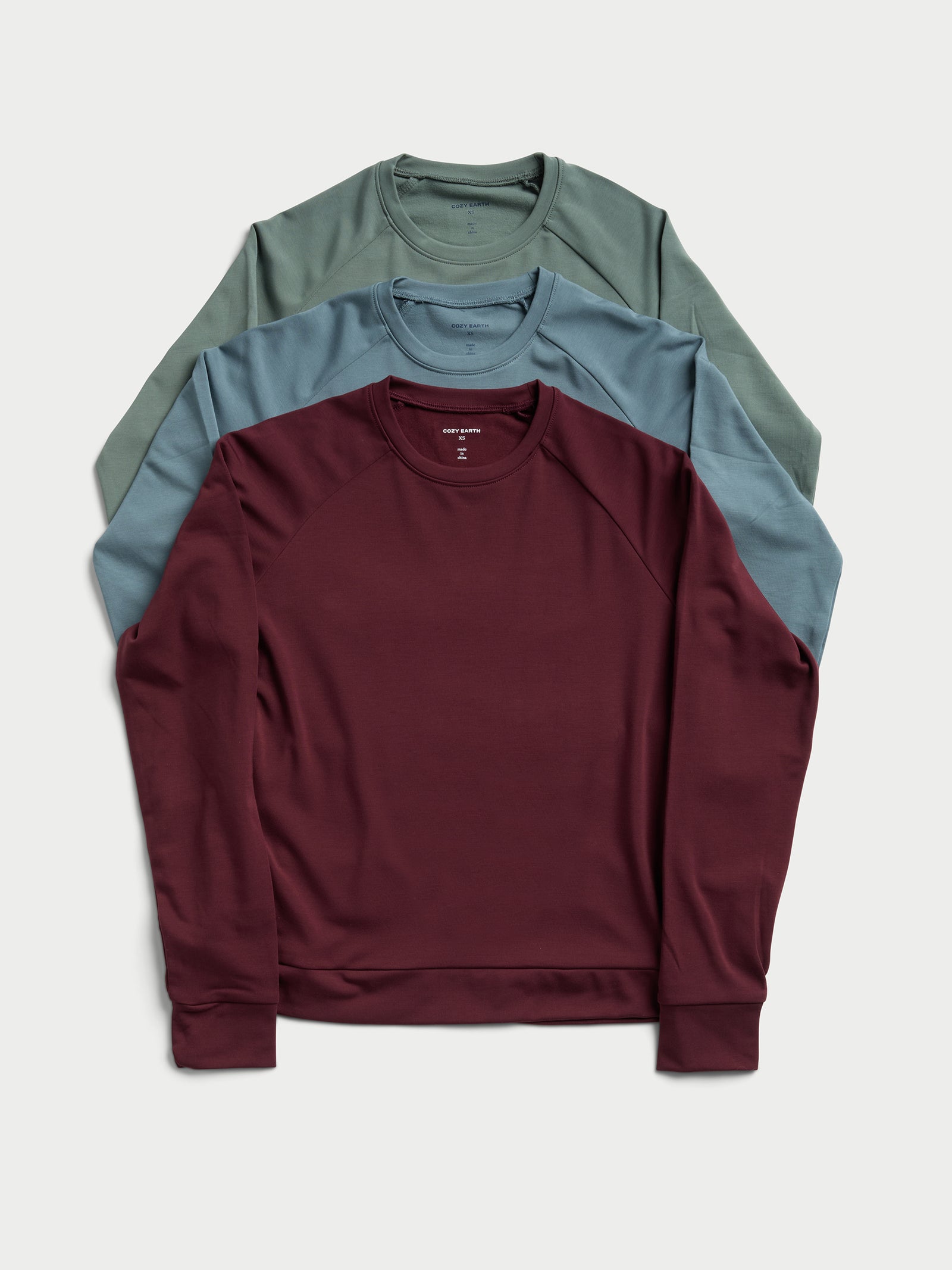 Three folded Women's Ultra-Soft Bamboo Pullover Crews by Cozy Earth are stacked. The top pullover is burgundy, the middle one is light blue, and the bottom one is sage green. They have a simple, casual design and are laid out on a plain light background. 