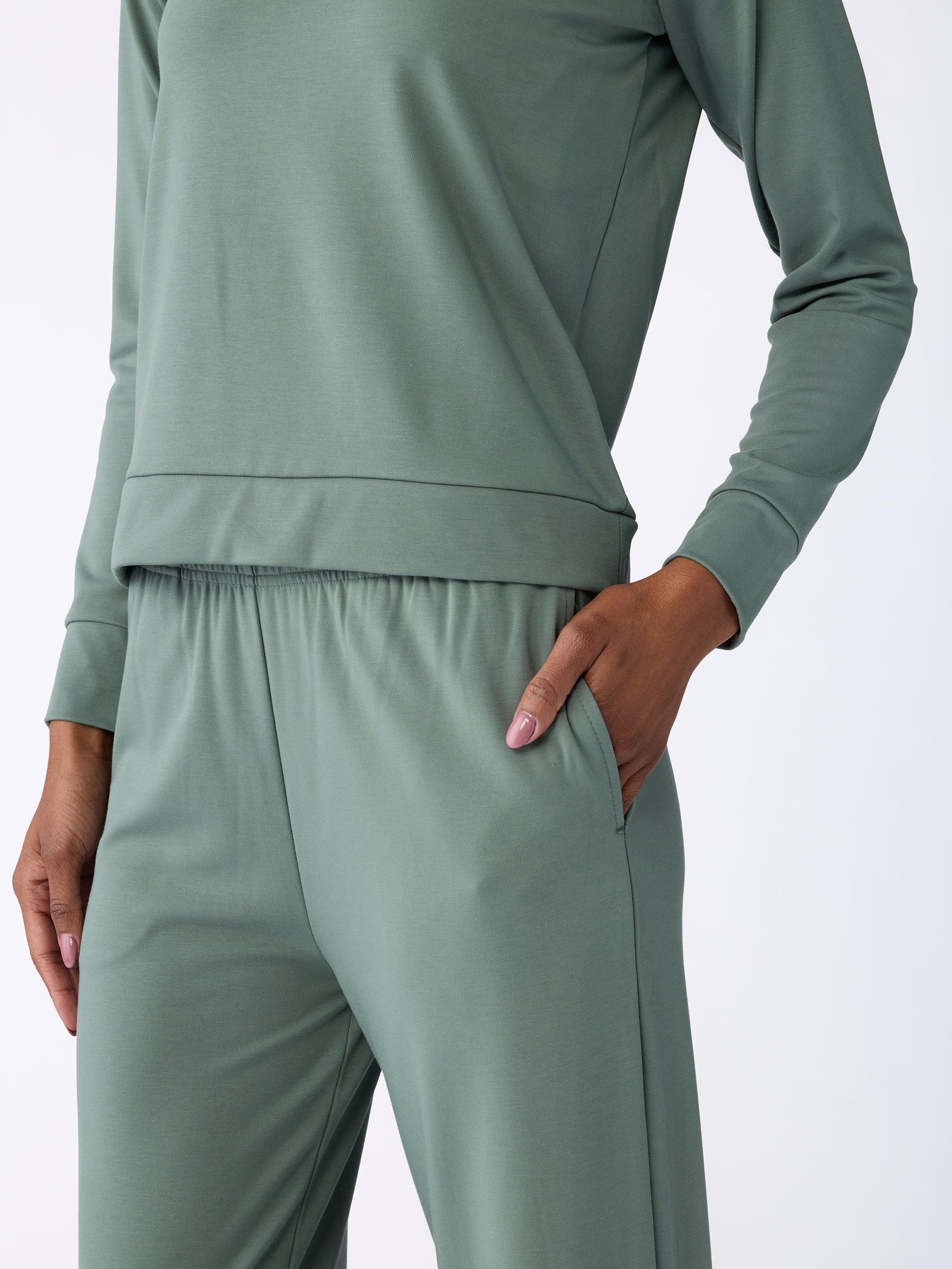 A person in Cozy Earth's Women's Ultra-Soft Bamboo Wide Leg Pull On Pant & Hoodie Set, sage green with a relaxed fit, has one hand in the pants pocket displaying a light pink manicure. The background is plain white. |Color:Oakmoss