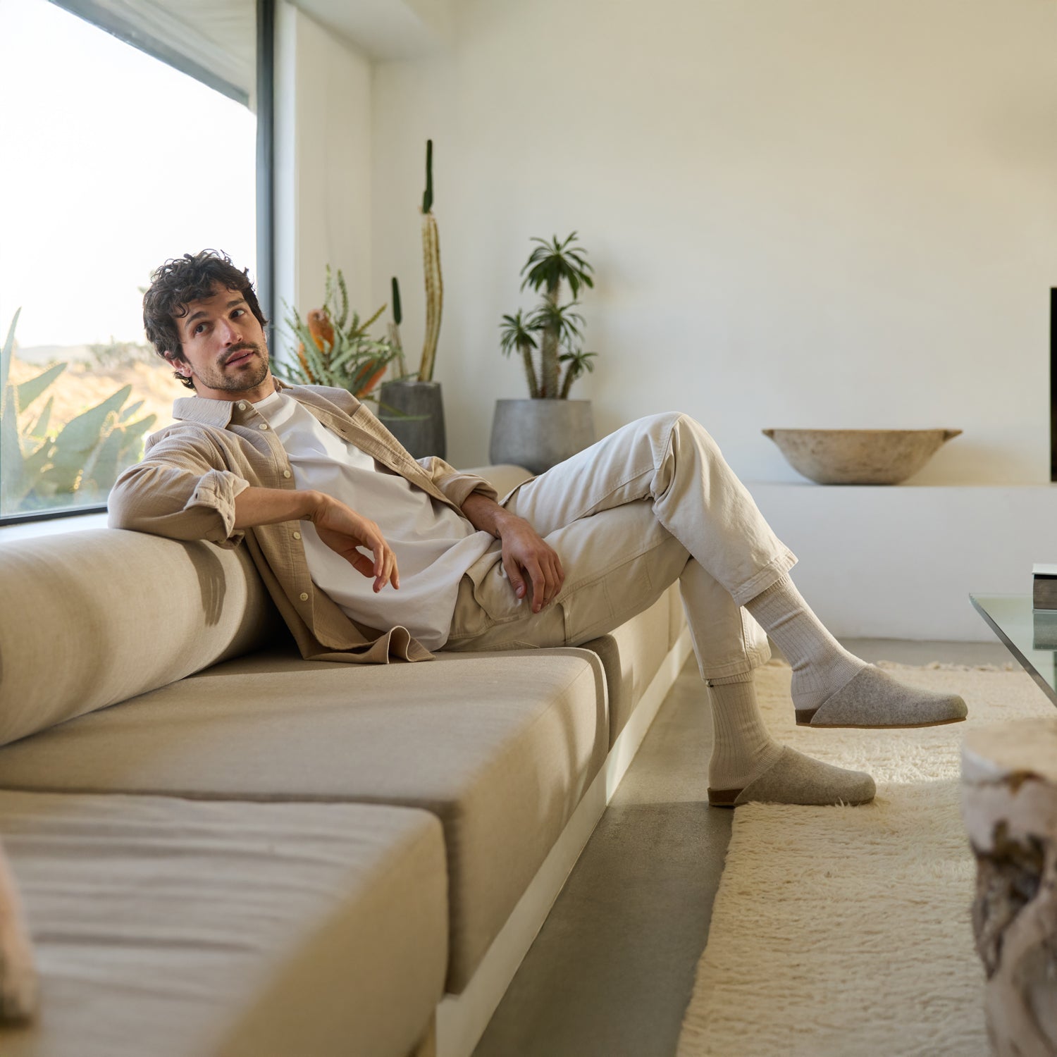 A person with curly hair sits relaxed on a beige sofa in a modern living room, wearing the light-colored Lakehouse Clog by Cozy Earth. Large windows flood the space with natural light, complemented by potted plants and minimalist decor. 