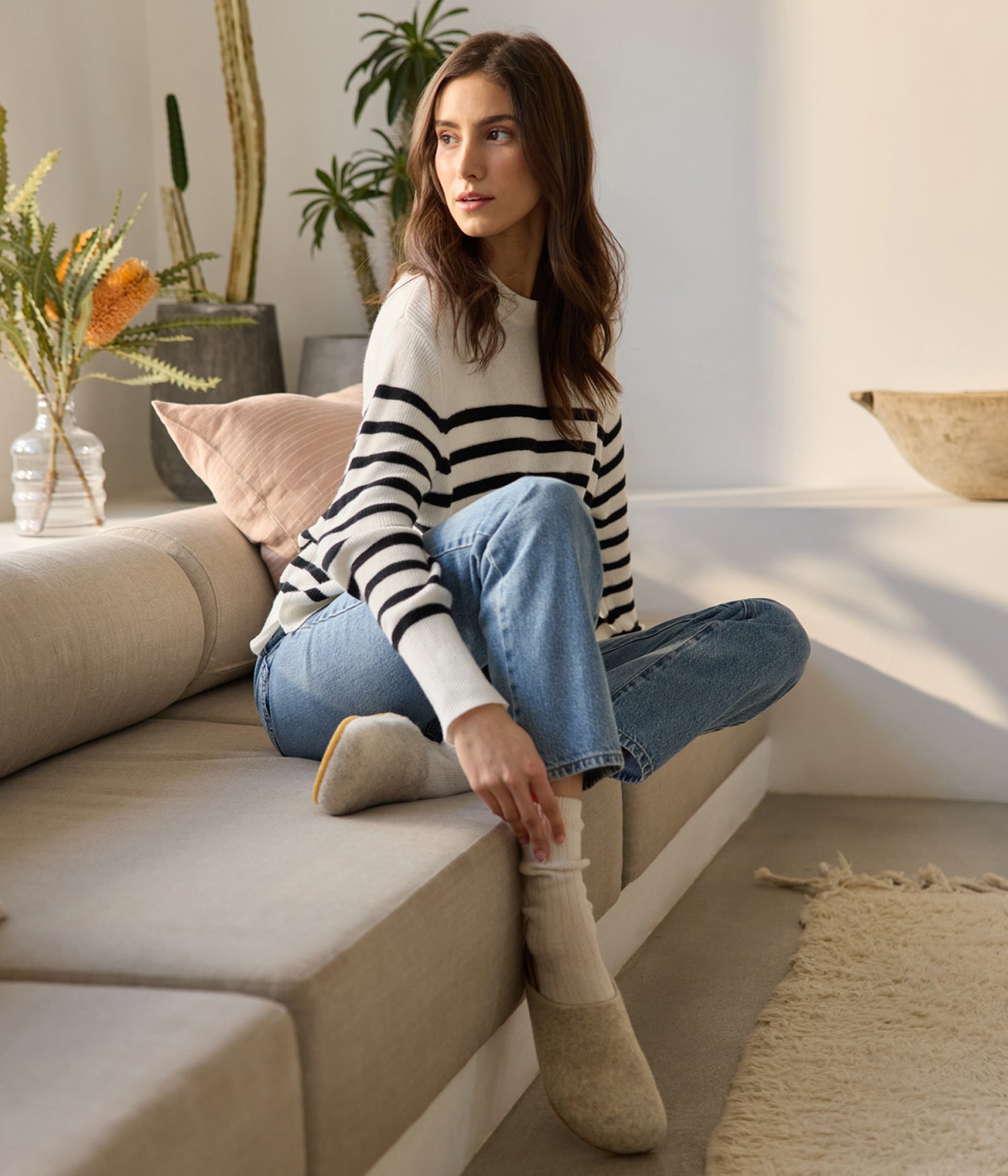 A woman lounges on a beige sofa, sporting a white and black striped sweater, blue jeans, and Lakehouse Clog boots by Cozy Earth. She gazes sideways as potted plants and a textured rug enhance the room's cozy ambiance. |Color:Oat