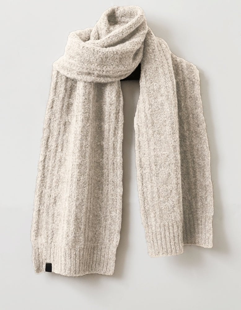 The Cozy Earth Cable Knit Scarf, in beige wool with a classic cable-knit pattern and ribbed ends, is displayed against a light gray background. Soft and cozy, it’s the perfect accessory for warmth and style. 