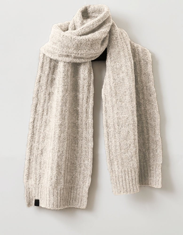 The Cozy Earth Cable Knit Scarf, in beige wool with a classic cable-knit pattern and ribbed ends, is displayed against a light gray background. Soft and cozy, it’s the perfect accessory for warmth and style. 