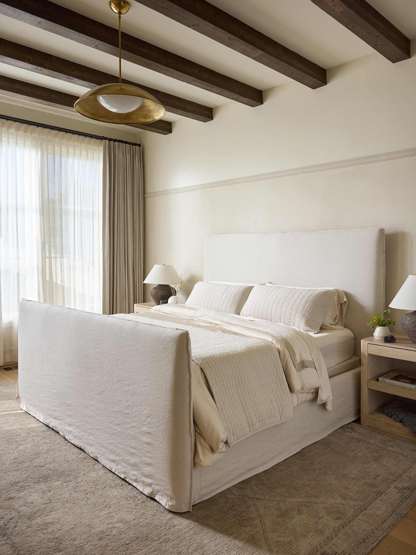 A tranquil bedroom showcasing a spacious bed adorned with a plush, light beige headboard and footboard, covered in white and beige bedding under a Cozy Earth Bamboo Blanket. The room features textured cream walls, wooden ceiling beams, a large window with sheer curtains, and bedside tables with white lamps. 