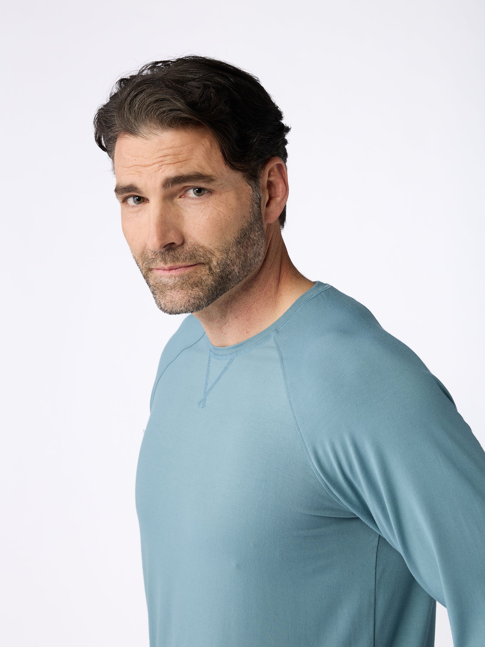 A man with dark hair and a beard is standing against a plain white background, wearing the Cozy Earth Men's Stretch-Knit Bamboo Long Sleeve in light blue, as he looks towards the camera with a neutral expression. 