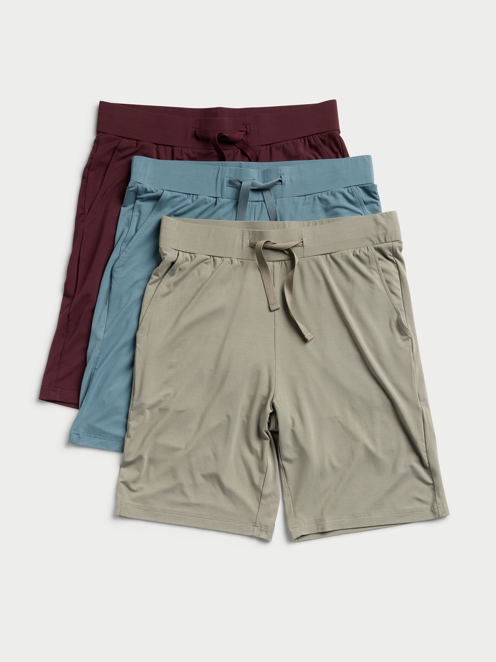 Three pairs of the Men's Stretch-Knit Bamboo Pajama Shorts by Cozy Earth are stacked on top of each other. The top pair is maroon, the middle one is light blue, and the bottom one is beige. Each pair features an elastic waistband with a drawstring and side pockets. 