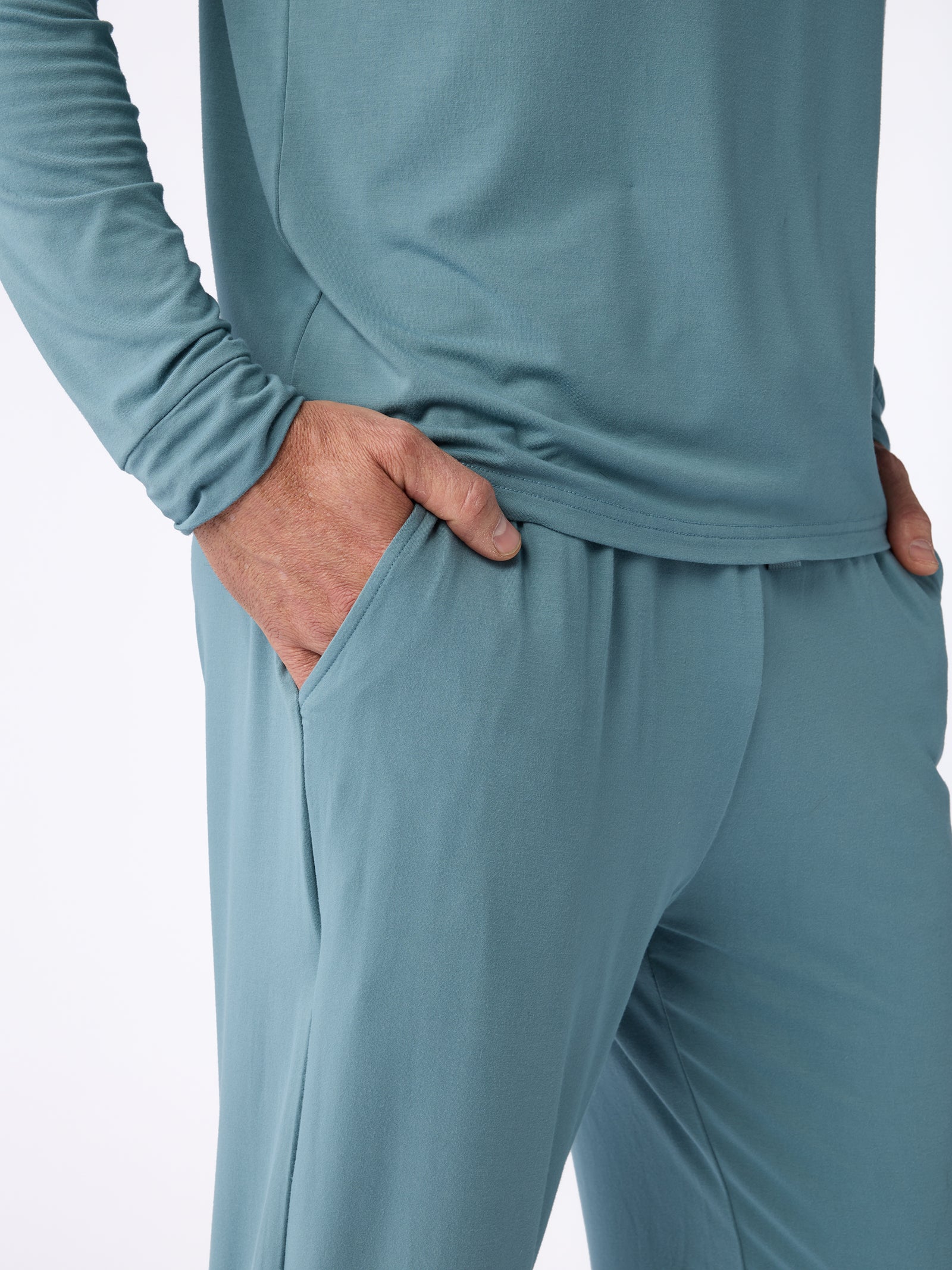 A person is wearing the Men's Stretch-Knit Bamboo Pajama Pant by Cozy Earth, shown in a teal outfit from shoulder to knee. They have on a long-sleeve shirt and matching pajama pants, with one hand in a pant pocket. The photo highlights the fabric's soft texture and comfortable fit. 