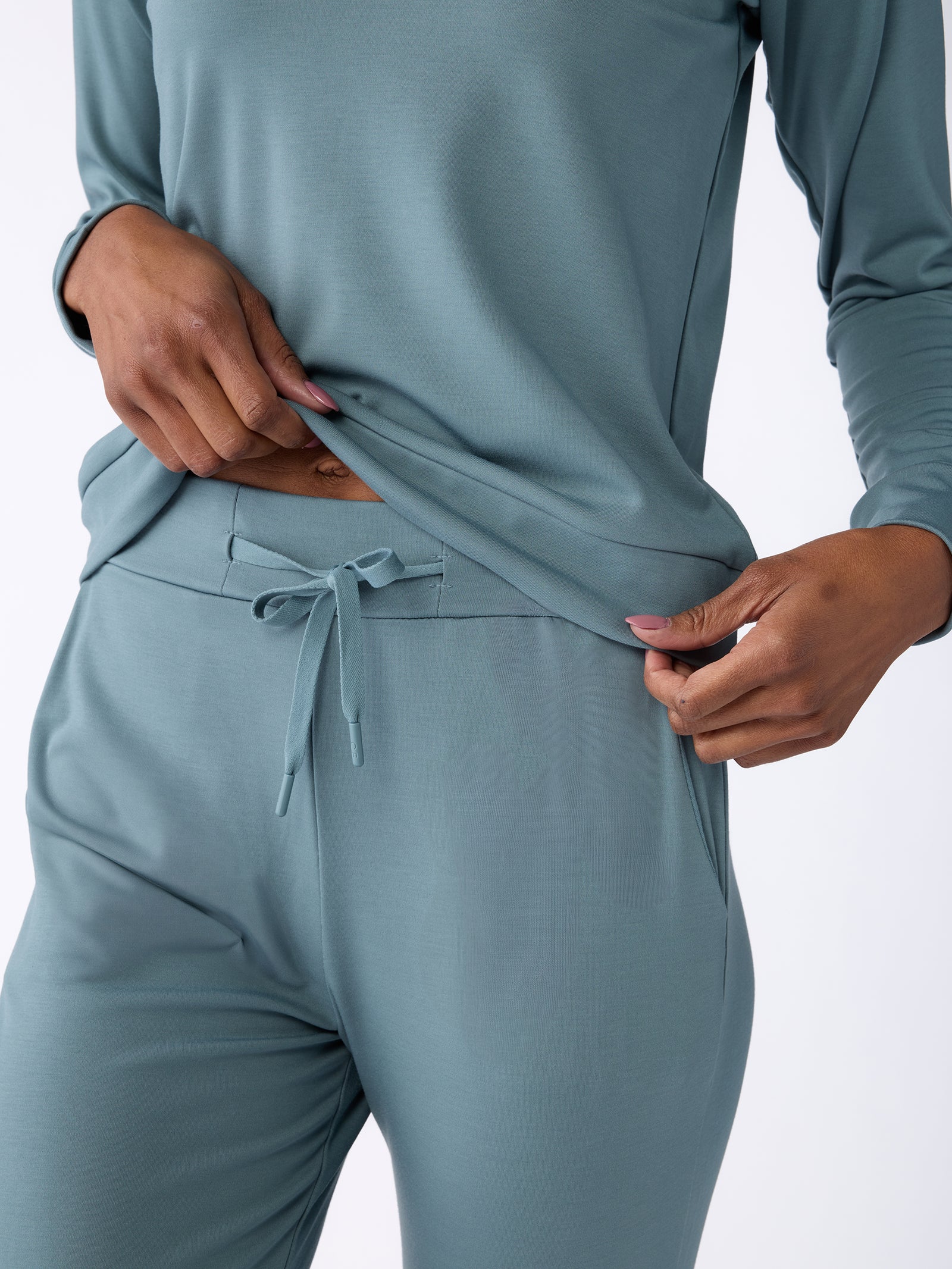 A person wearing the Women's Bamboo Jogger Pant by Cozy Earth, featuring a light blue, long-sleeved top and matching pants with a drawstring waist, gently pulling the shirt. The clothing highlights the soft fabric and relaxed fit. 