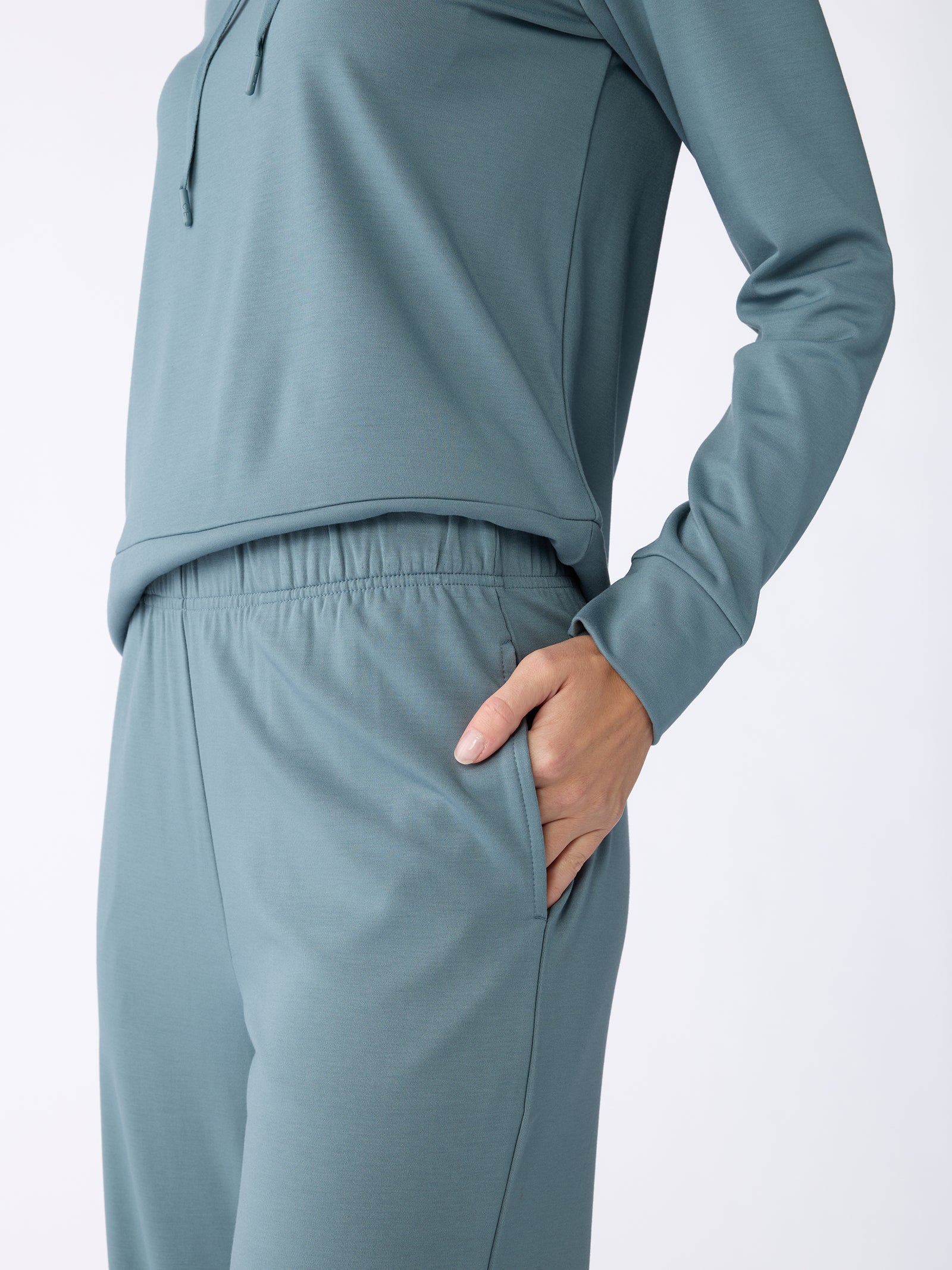 A person wearing a teal Cozy Earth tracksuit with their hand in the right pocket, showcasing a casual outfit that includes the Women's Ultra-Soft Bamboo Wide Leg Pull On Pant, complemented by a long-sleeved top, set against a plain white background. 