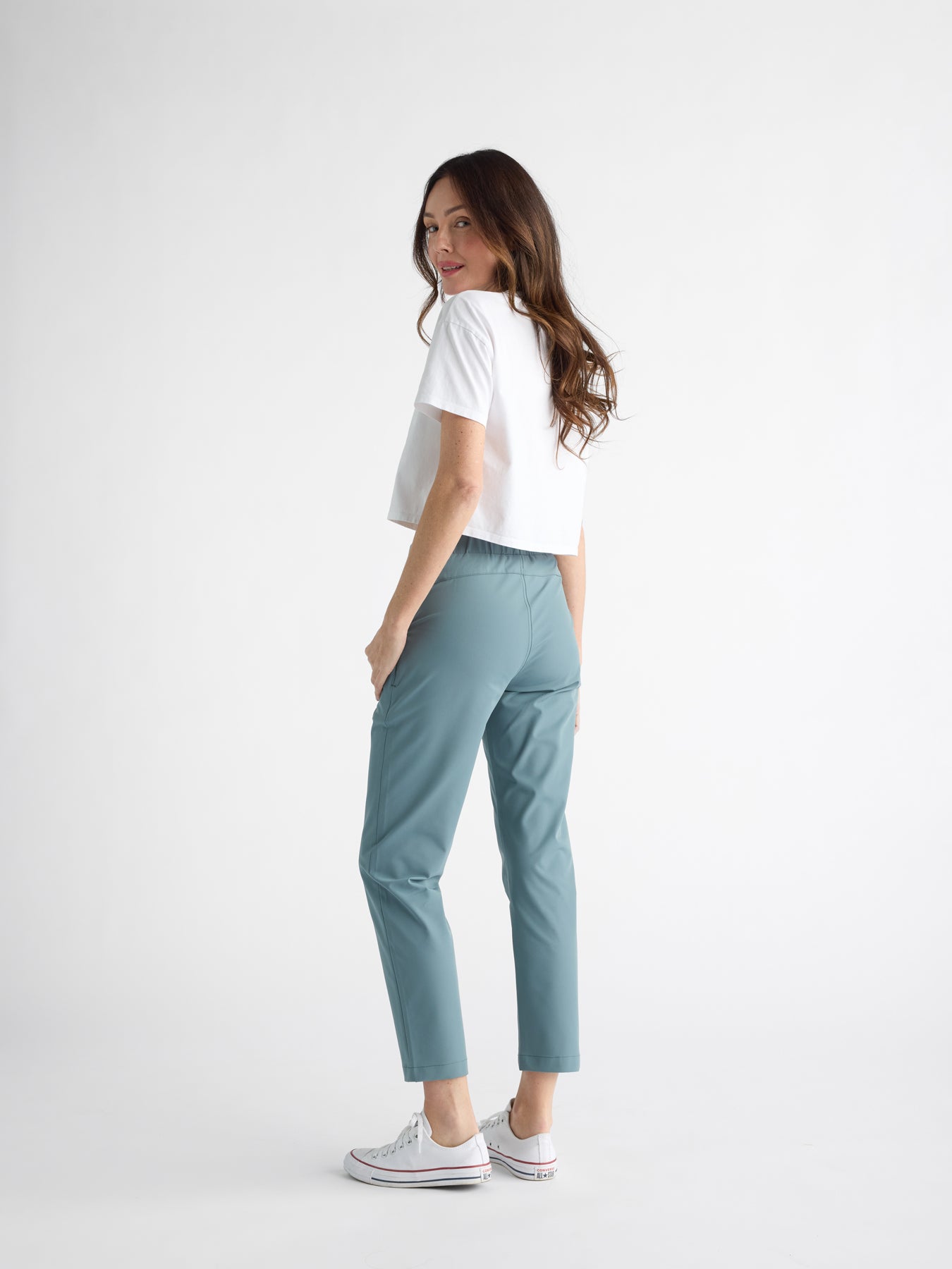 A brown-haired person wears a white short-sleeve shirt, Cozy Earth's Women's Always Cropped Pant in teal, and white sneakers. They stand against a plain white background, glancing over their shoulder. 