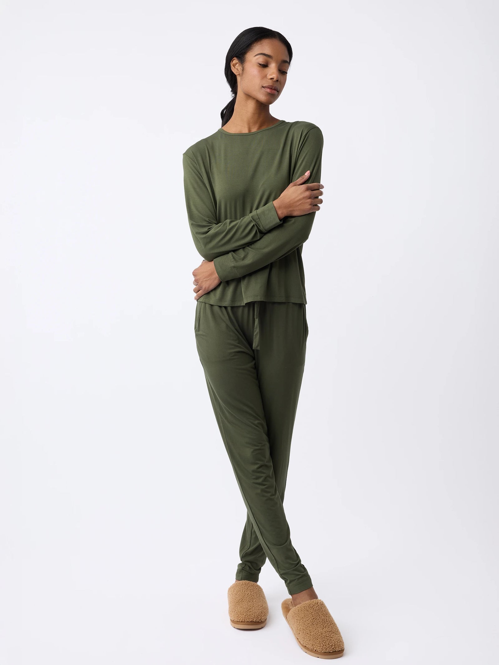 A woman wears a matching dark green Women's Stretch Knit Bamboo Long Sleeve Lounge Tee and pants with arms crossed over their chest. They pair it with light brown slippers, standing against a plain light-colored background. The outfit is by Cozy Earth. 