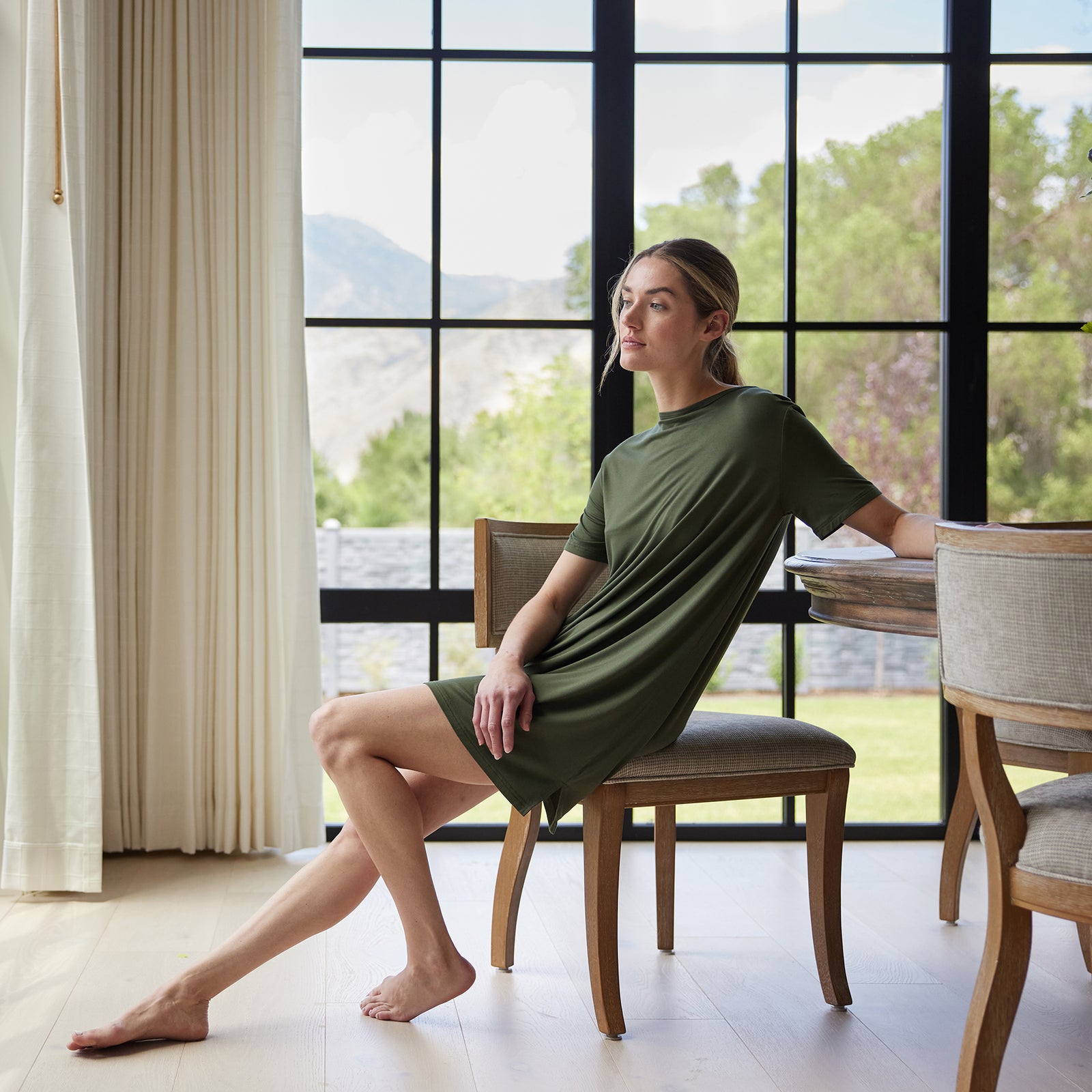 A woman in a Women's Bamboo Stretch Knit Sleep Dress from Cozy Earth sits casually on a wooden chair near a dining table, positioned by a large window with a scenic view of trees and mountains. She has one leg up on the chair and appears relaxed, with sheer curtains partially drawn on one side. 