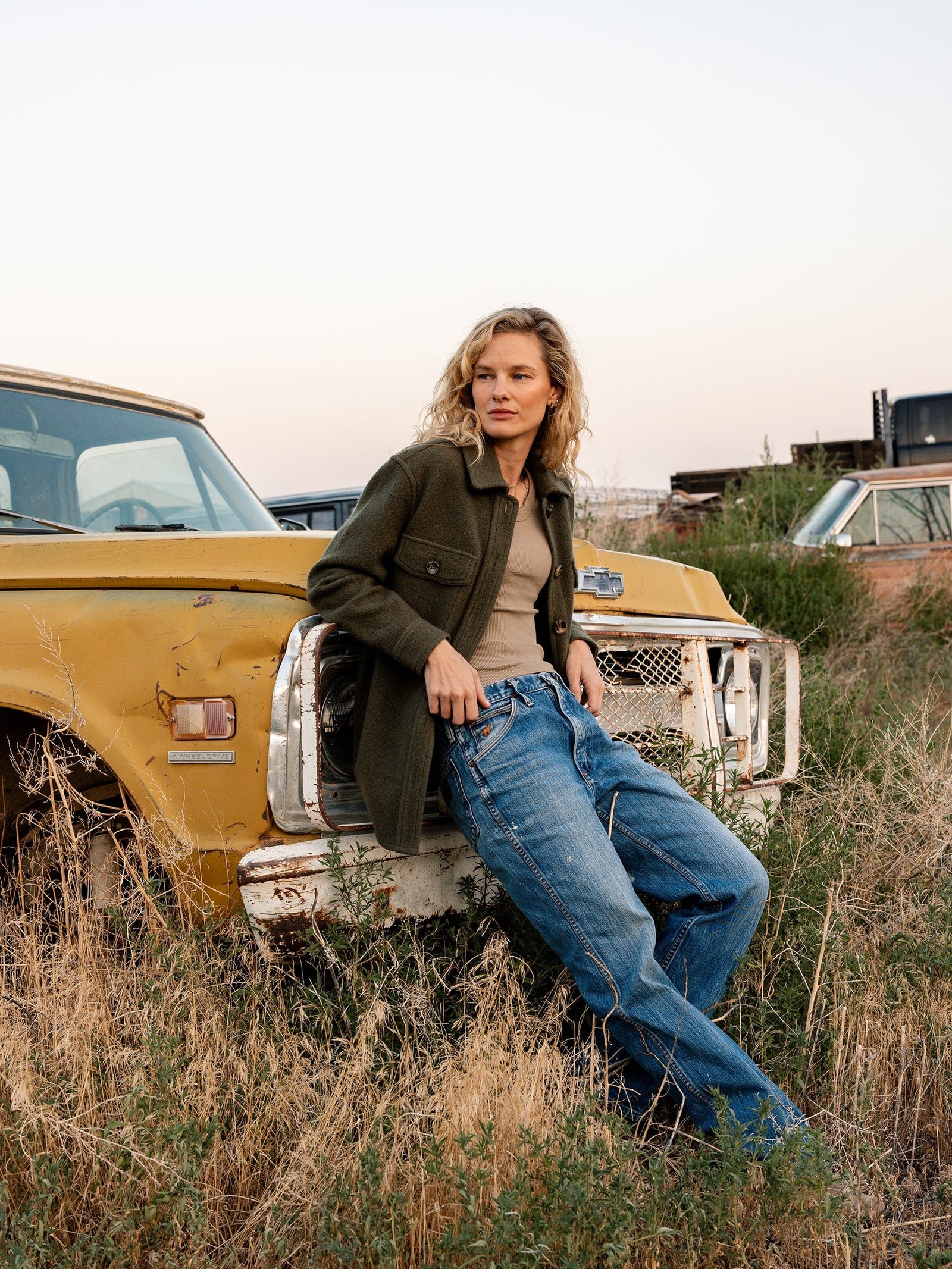 A person with wavy blonde hair leans against a yellow, rusty vintage truck. They are wearing Cozy Earth's Women's Boucle Shacket, a beige top, and blue jeans. The background includes tall grass, an open field, and more old vehicles under a cloudy sky. 